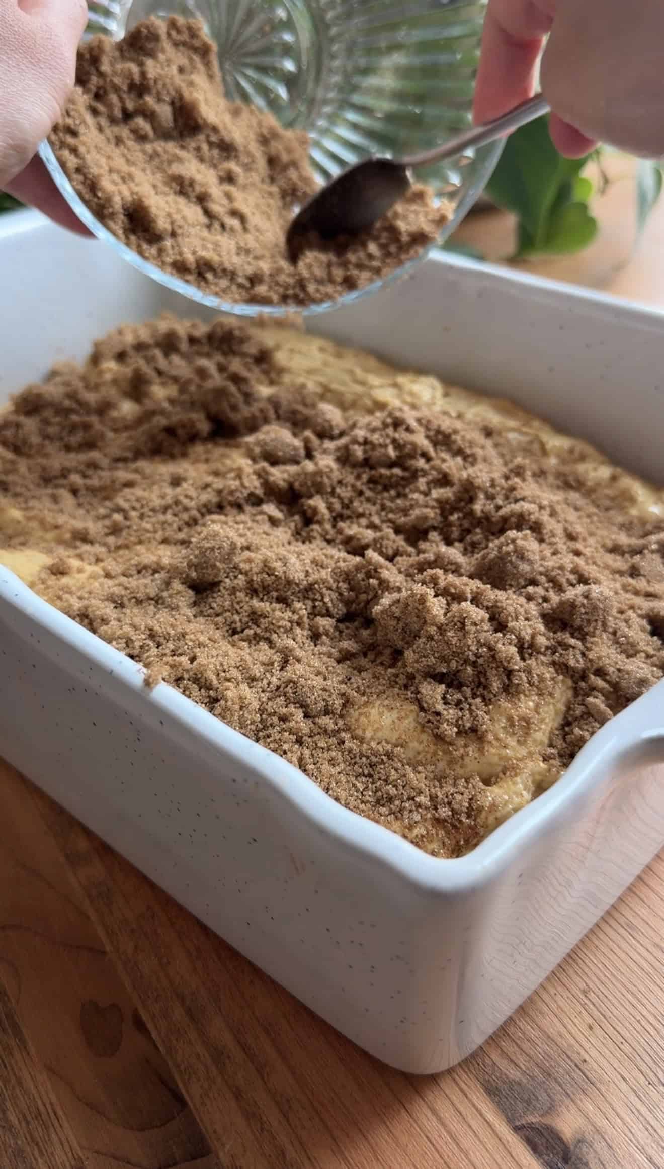 Brown sugar being sprinkled from a glass bowl onto focaccia dough in a ceramic baking dish.