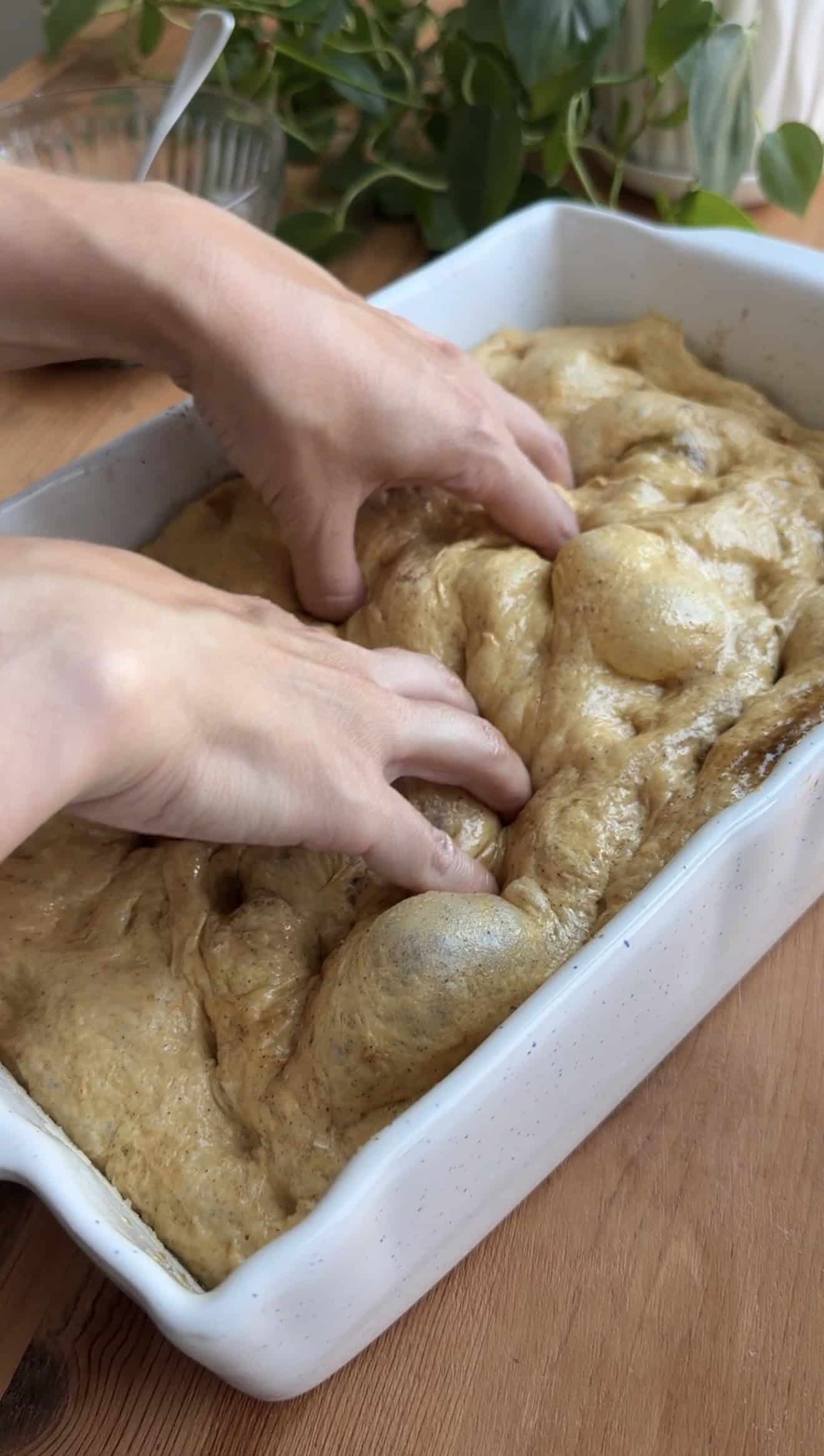 Two hands dimpling bubbly focaccia dough in a light-colored ceramic baking dish.