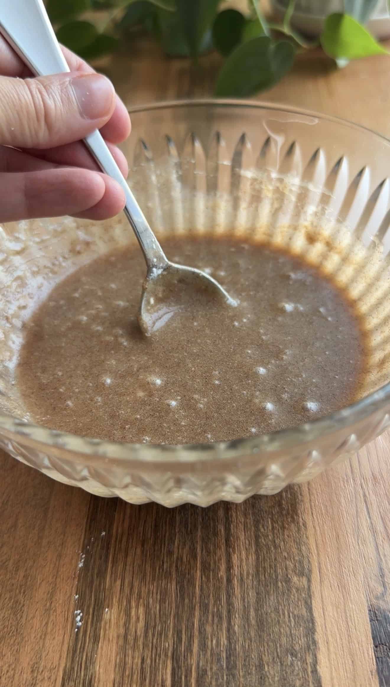 A hand stirring thick, brown glaze in a glass bowl with a metal spoon.