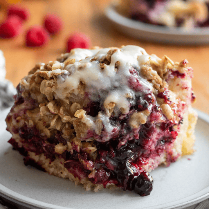 A large square of sweet focaccia filled with blueberries and raspberries and topped with oats and a white glaze sitting on a grey plate.