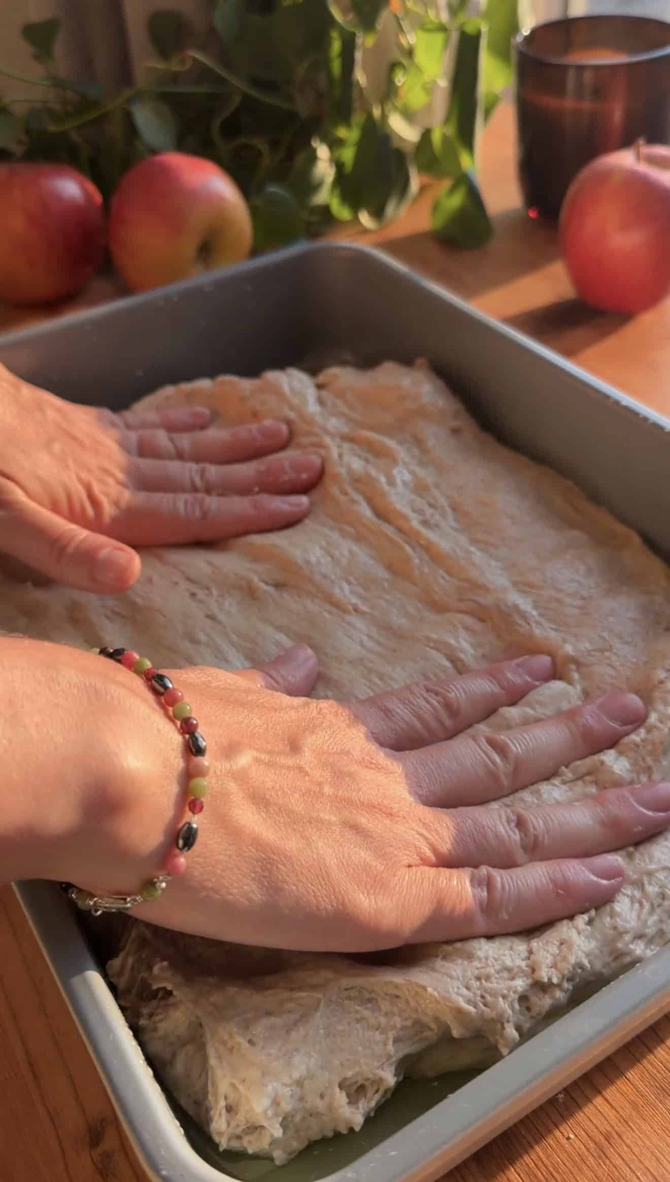 Two hands pressing focaccia dough into a greased metal baking pan.