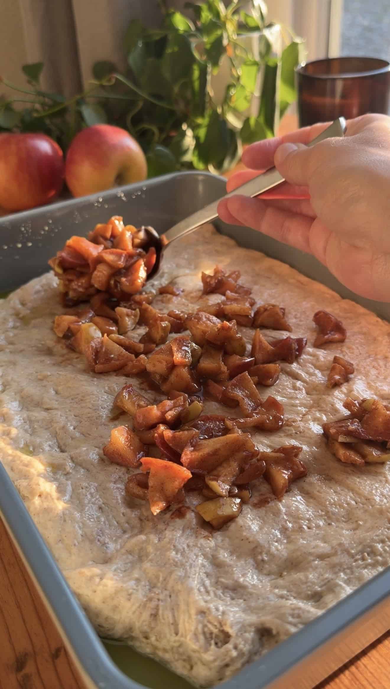 A hand spooning cooked, diced apples onto focaccia dough spread out in a baking dish.