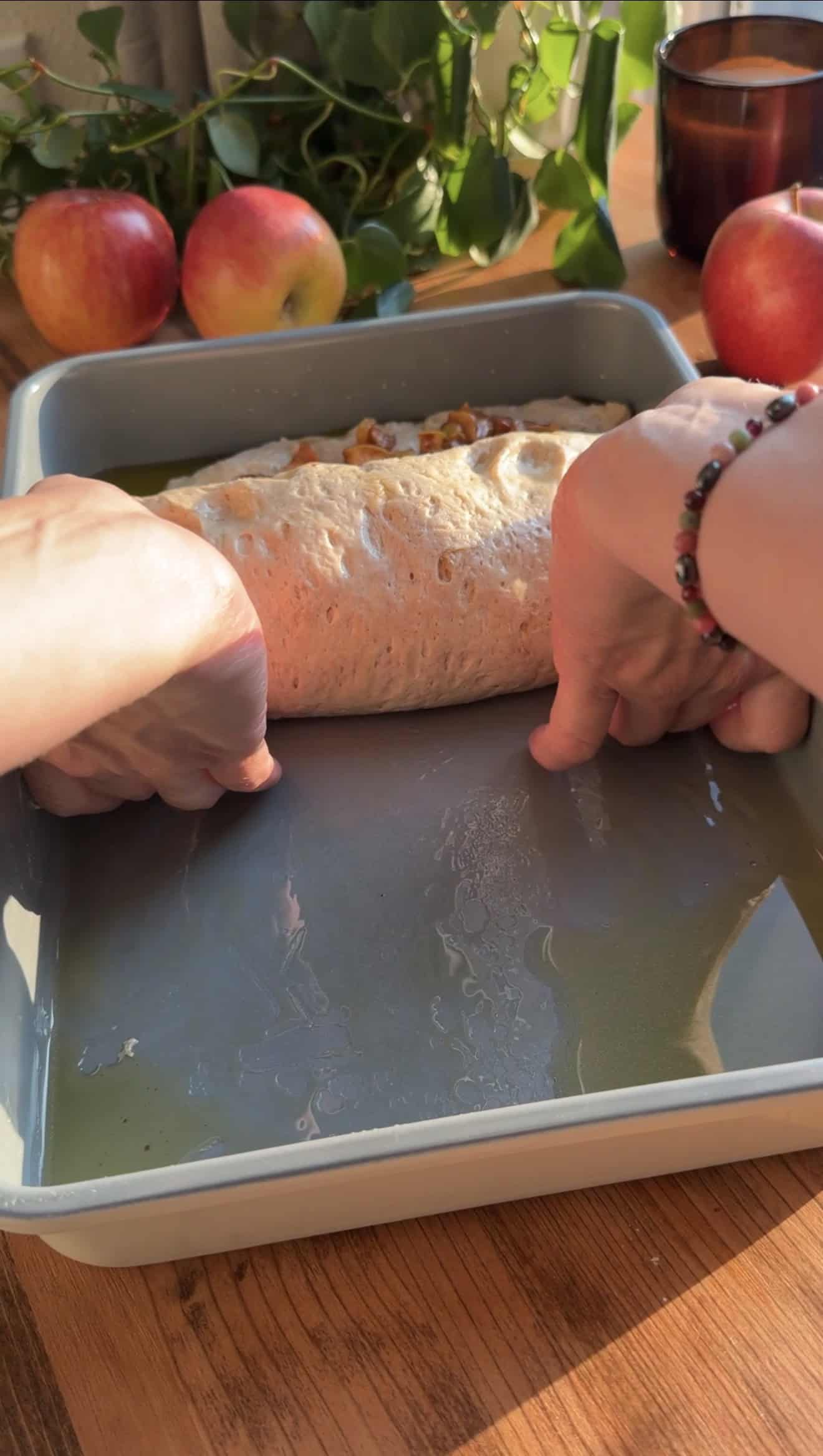 Two hands rolling up diced, cooked apples into focaccia dough inside a metal baking dish on a table.