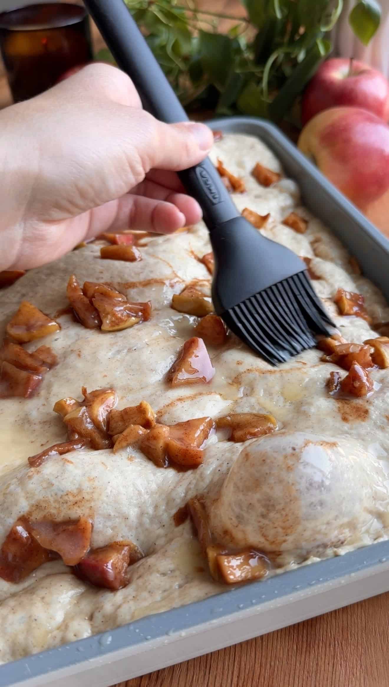 A hand brushed melted butter onto proofed, bubbly focaccia dough topped with small pieces of cooked cinnamon apples.