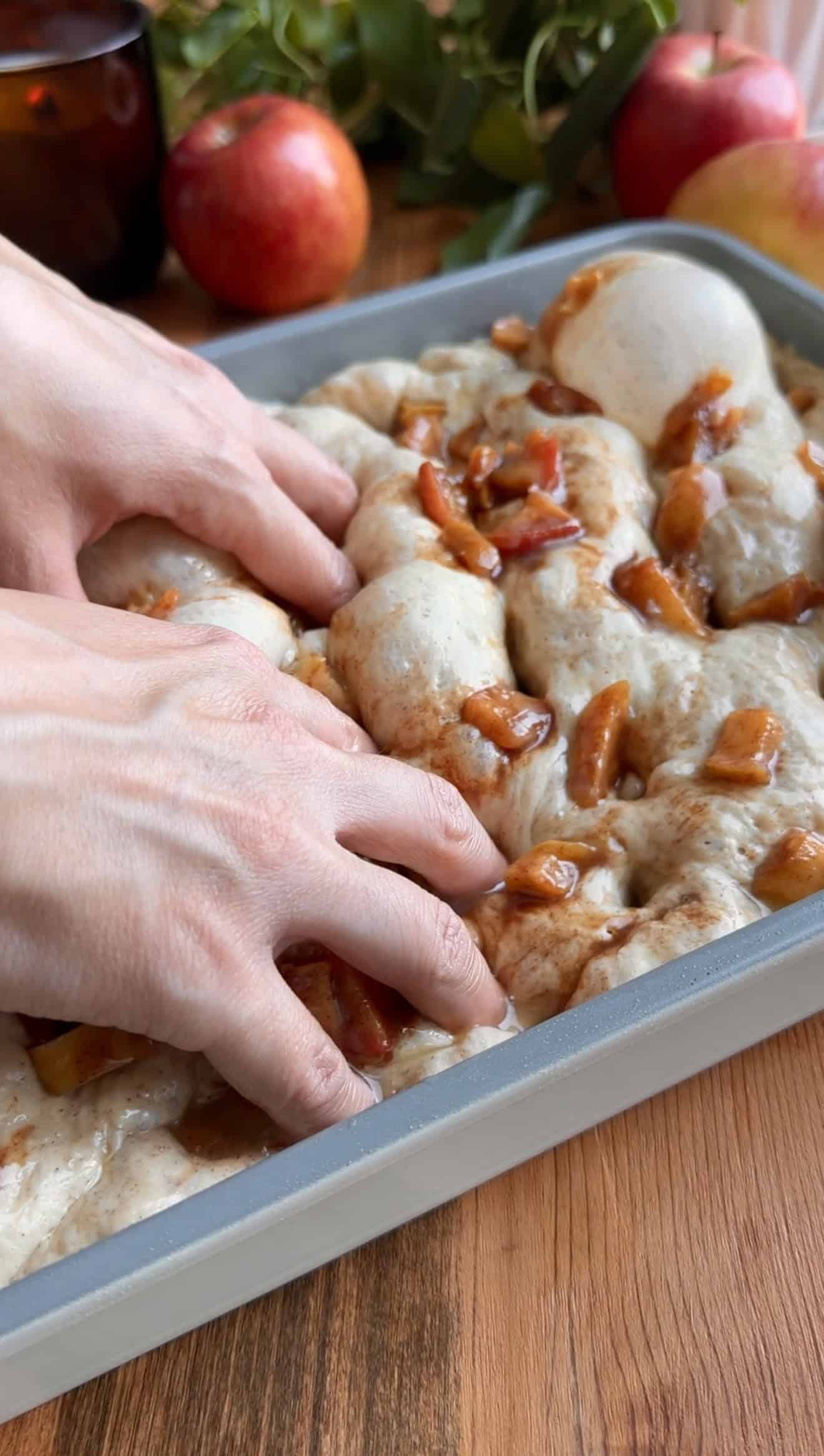 Two hands dimpling focaccia dough in a metal baking dish, with the dough topped with diced cinnamon-coated apples.