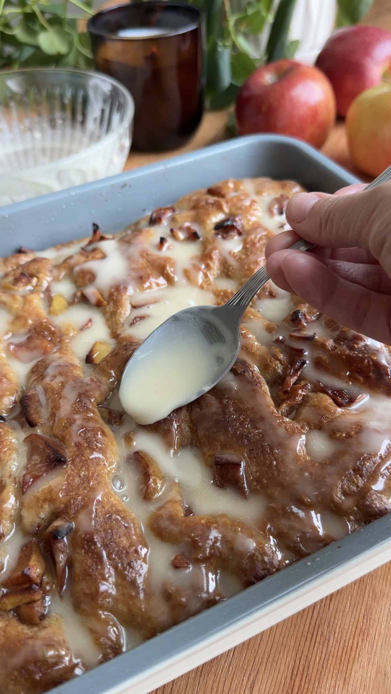 A hand drizzling glaze onto baked apple fritter focaccia in a metal baking dish.