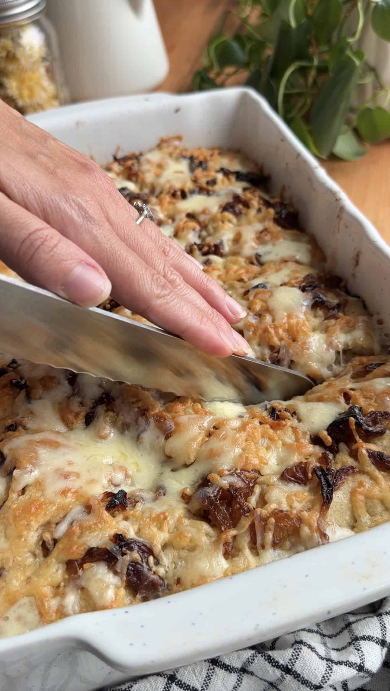 Someone slicing cheesy onion focaccia with a serrated knife in a white casserole dish.