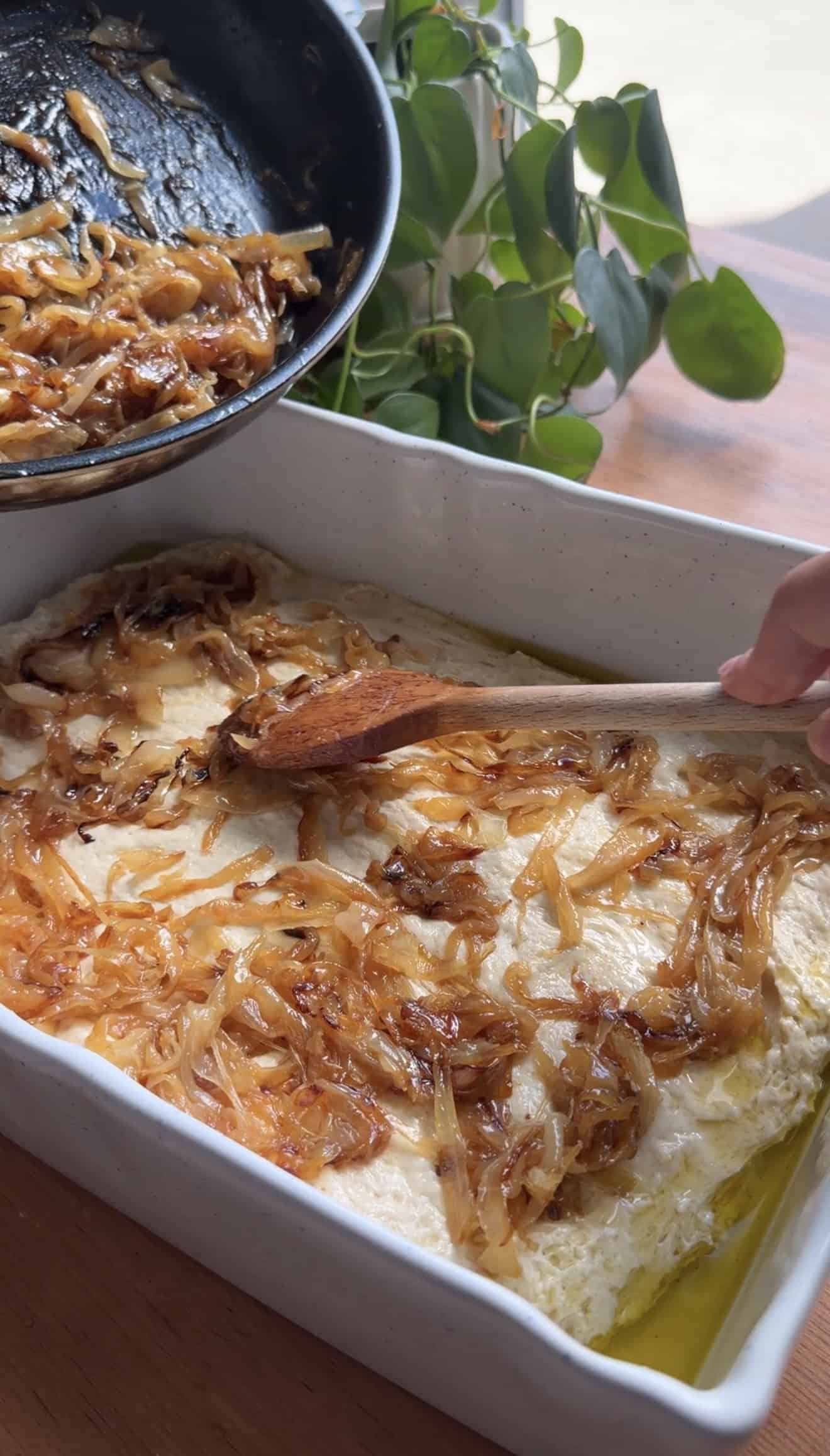 A hand spooning caramelized onions from a saucepan onto focaccia dough spread in a baking dish.