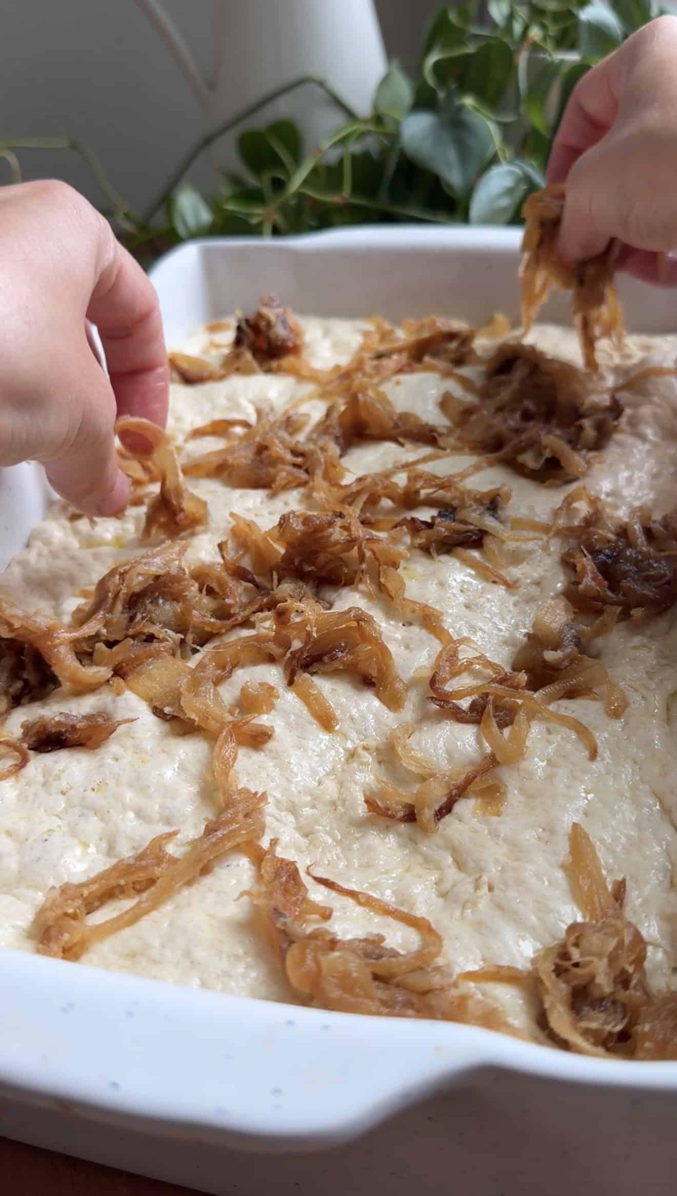 Two hands spreading caramelized onions on top of bubbly, risen focaccia dough in a baking dish.