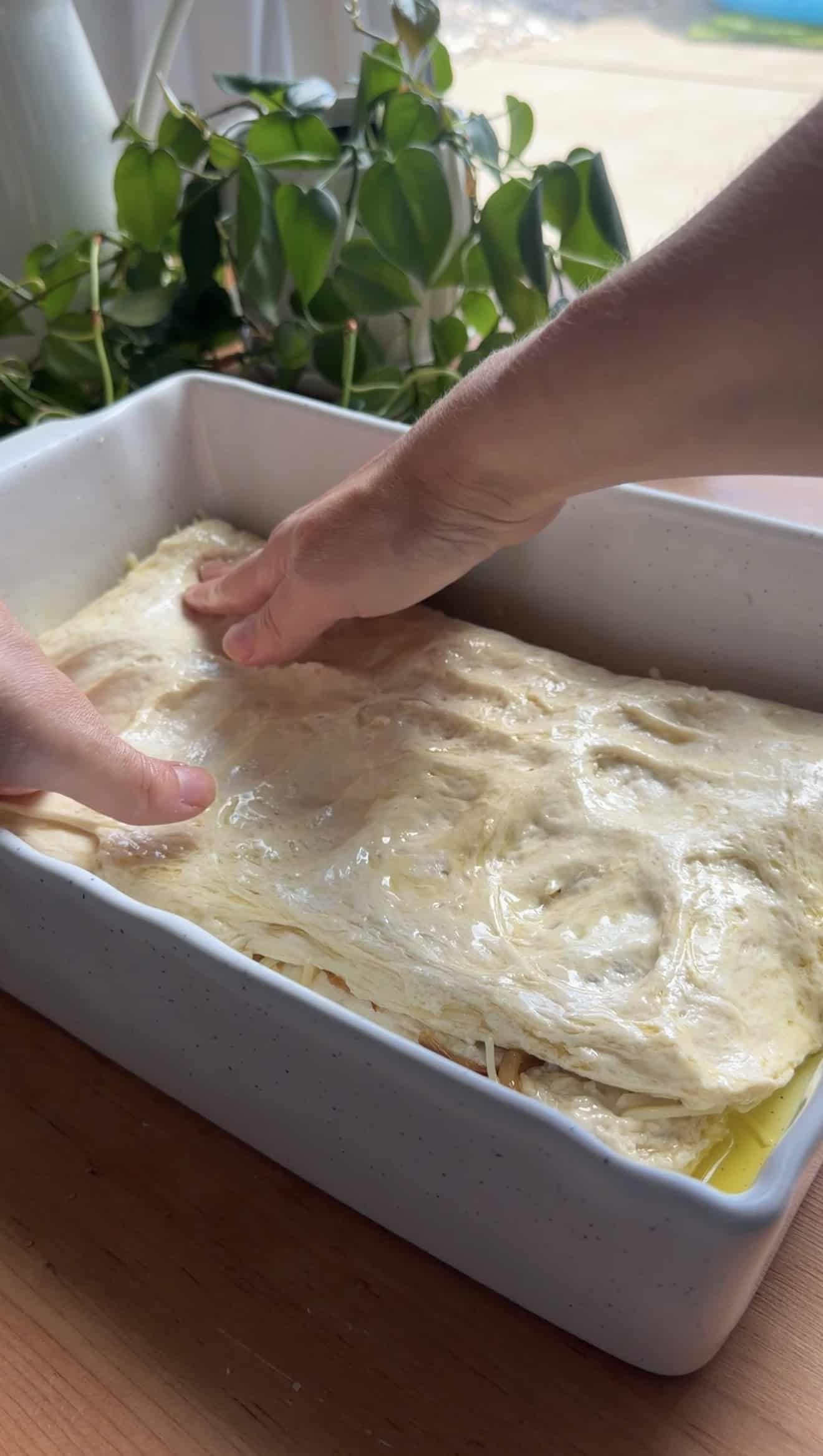 Two hands spreading focaccia dough in a white baking dish.