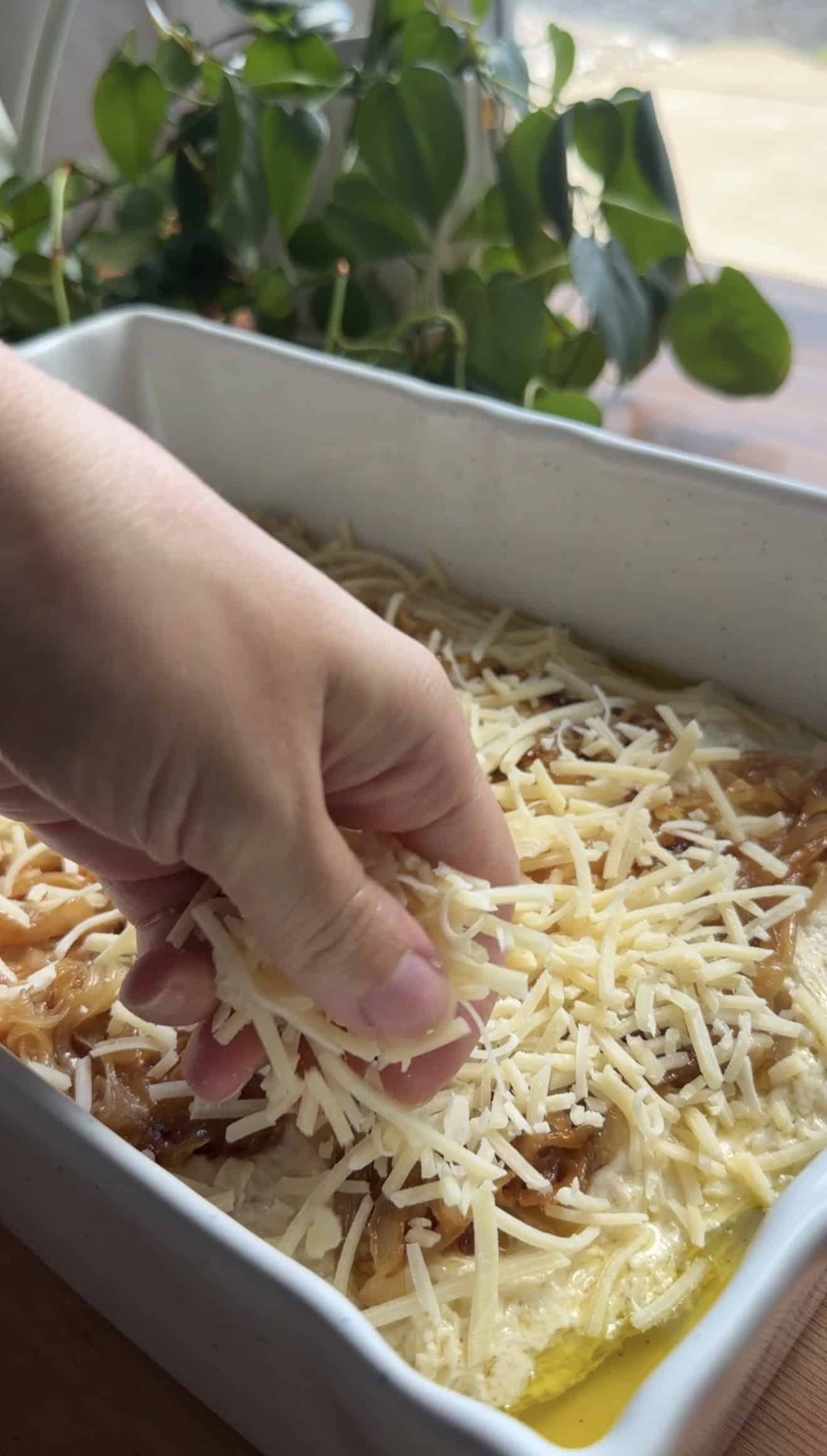 A hand sprinkling shredded cheese onto focaccia dough topped with caramelized sliced onions.