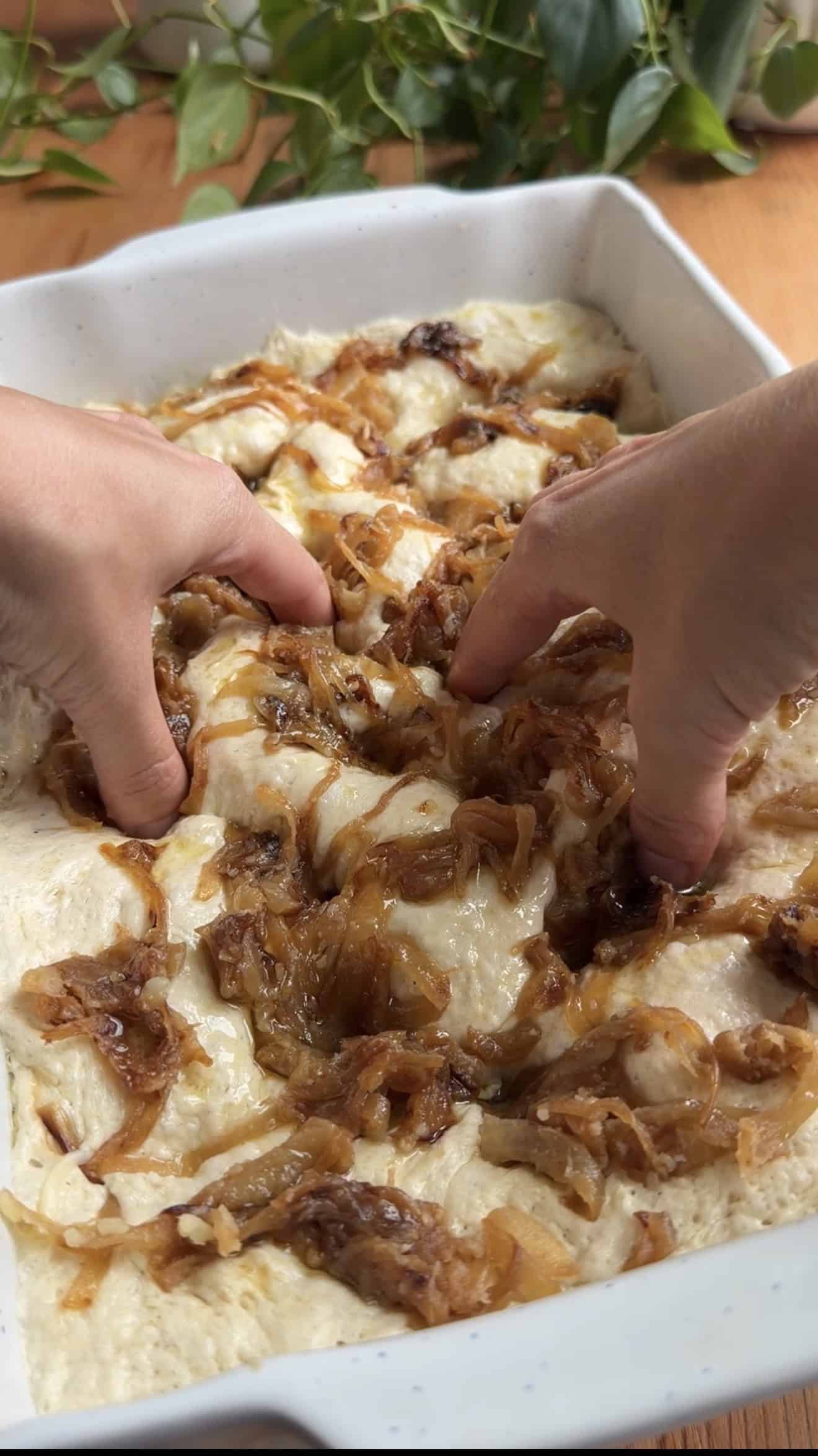 Two hands indenting bubbly focaccia dough topped with golden, caramelized onions.
