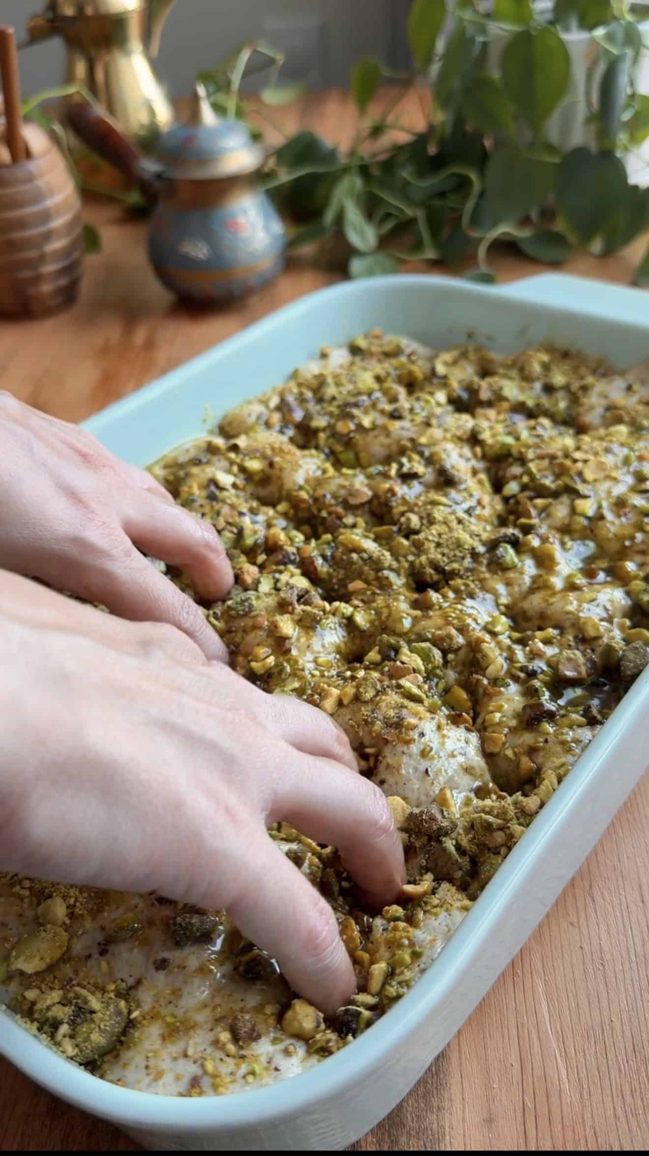Two hands dimpling indentations into focaccia dough topped with pistachios in a baking dish.