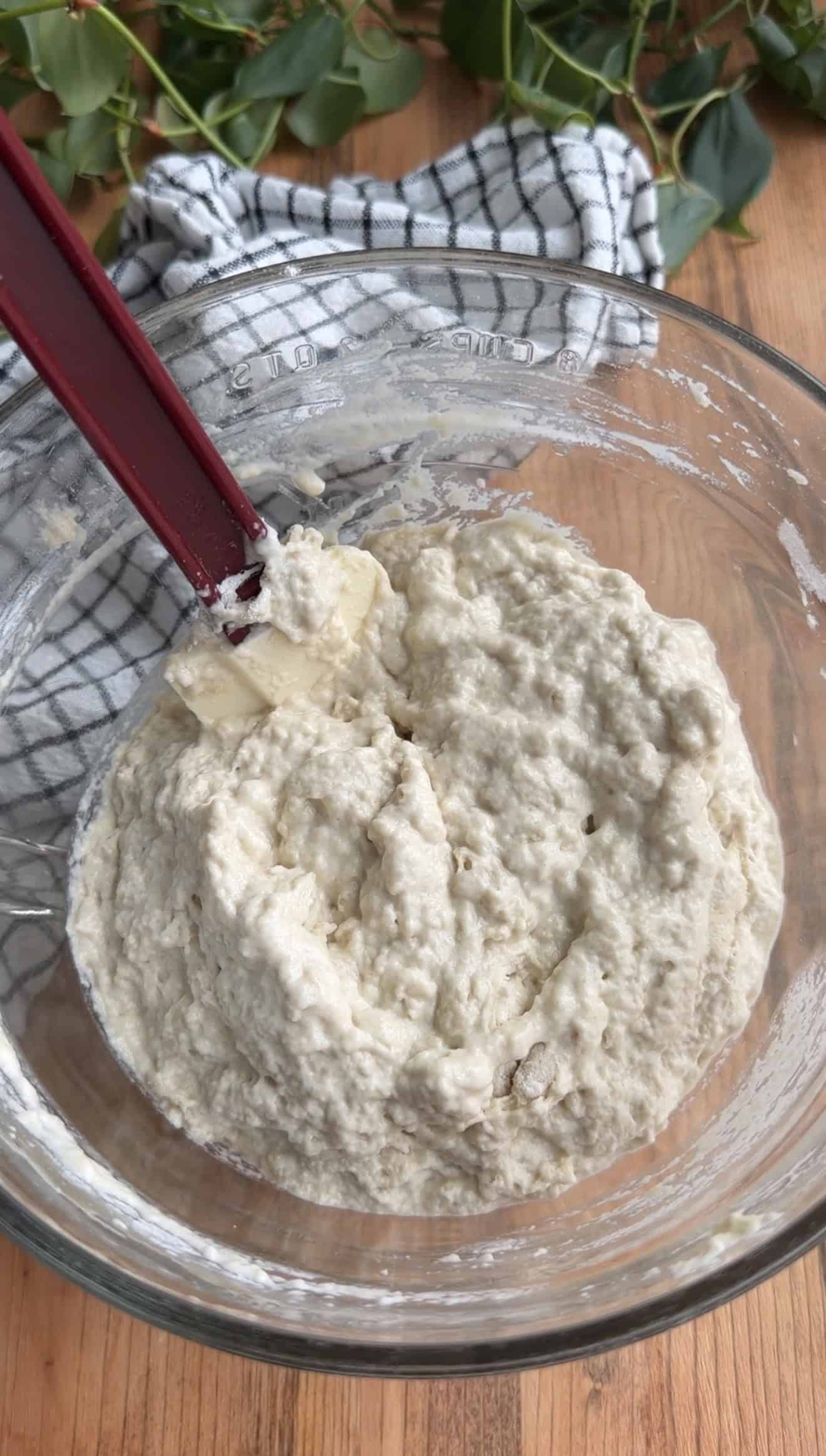 A sticky bread dough in a glass mixing bowl with a silicone spatula sitting in it.