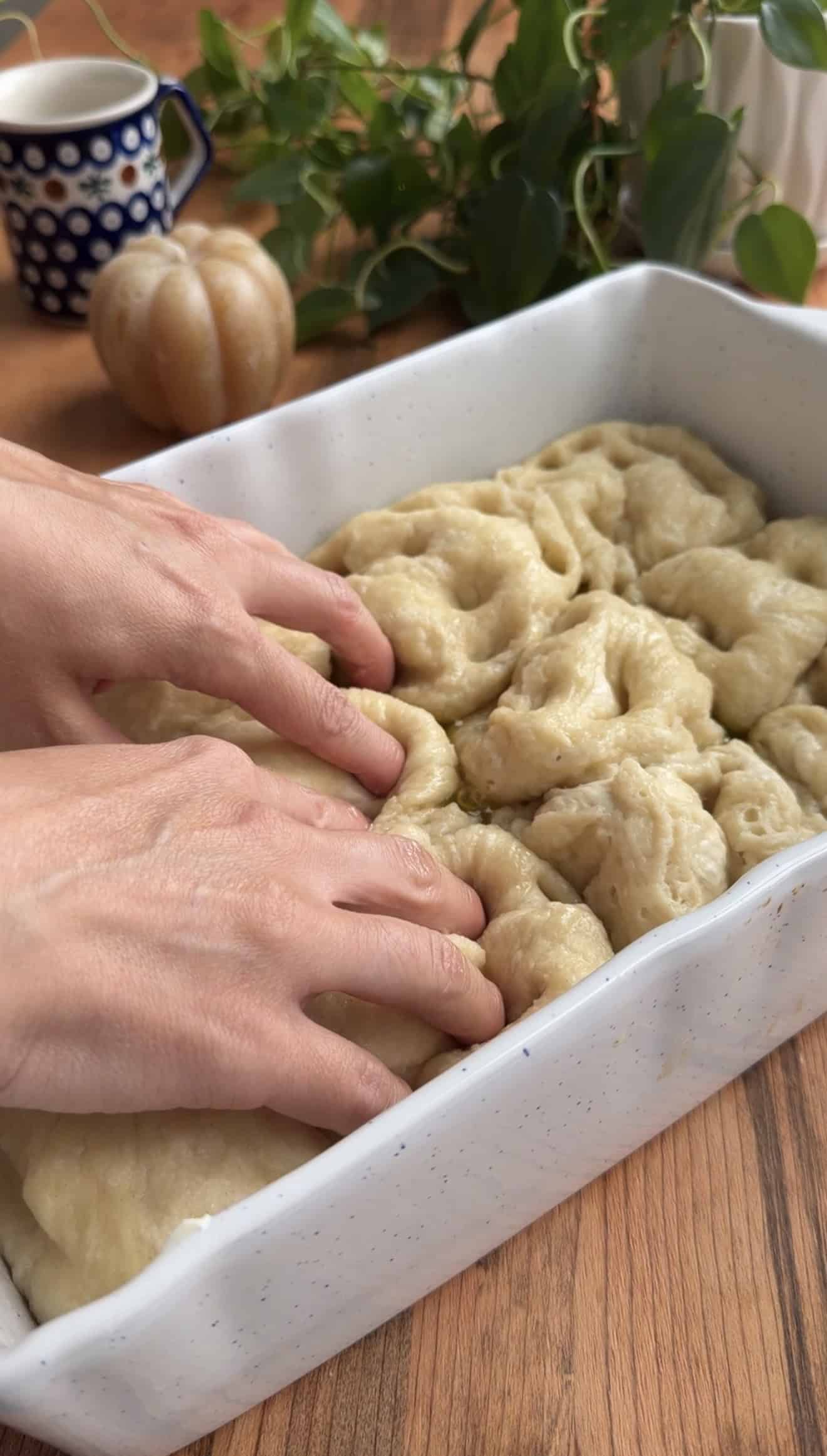 Two hands pressing dimples into focaccia dough in a white baking dish on a wooden table.