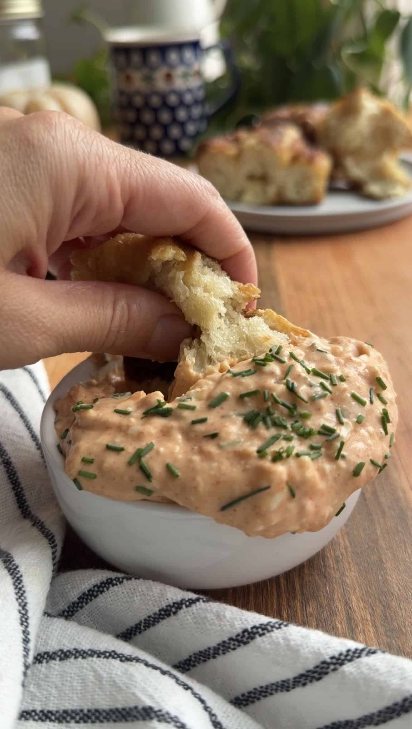 A hand dipping a torn piece of pretzel focaccia into a small white bowl overflowing with cheese dip and chives.