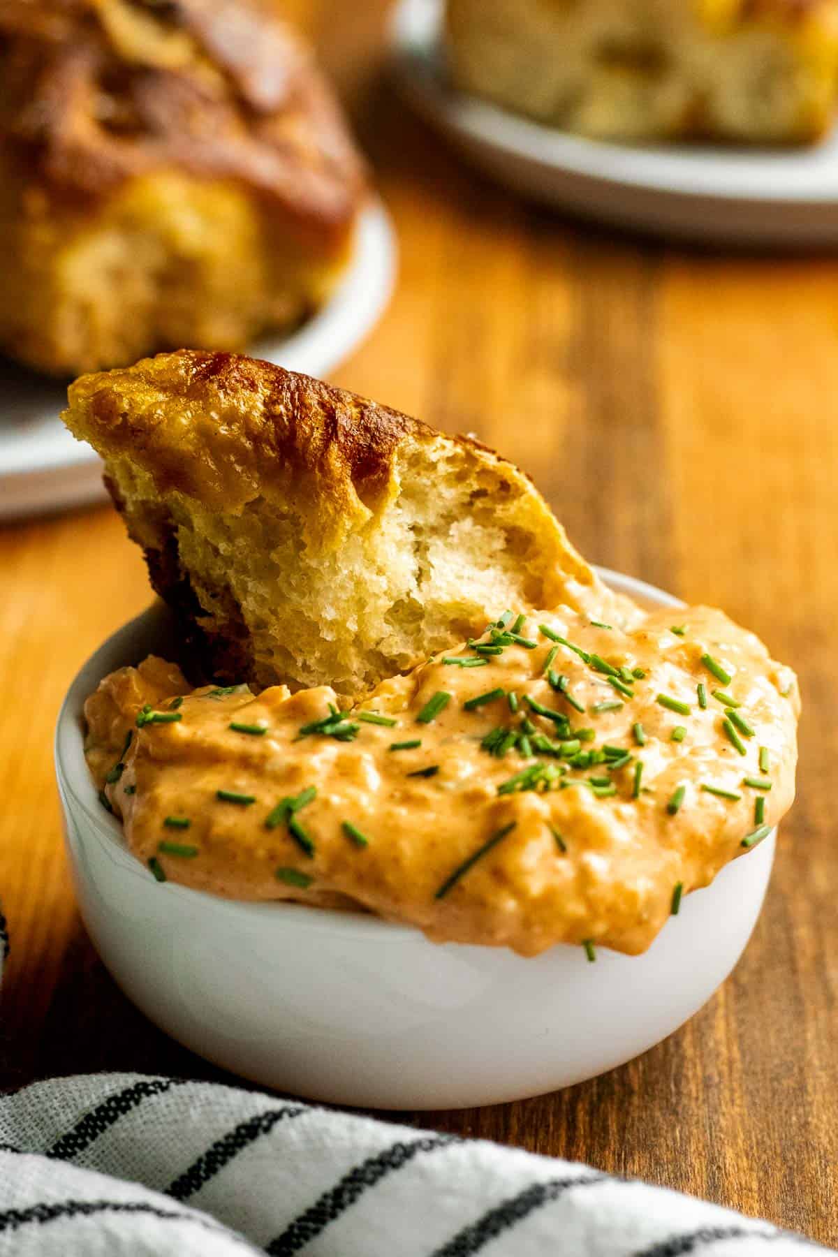 A piece of pretzel focaccia sitting in a bowl of chunky cheese dip topped with chopped fresh chives.