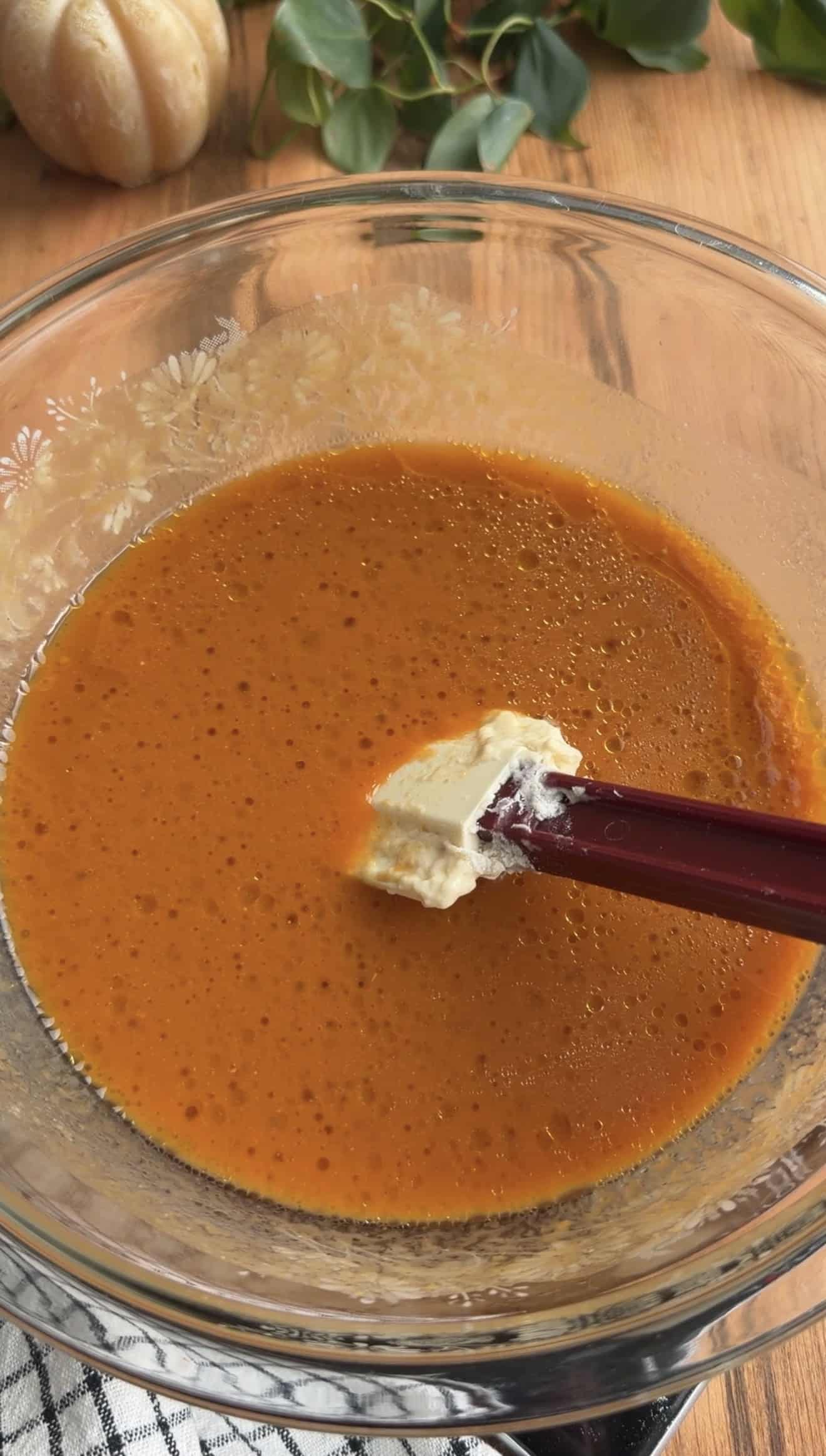 A bowl of a thing orange liquid with a silicone spatula sitting on a wood table.