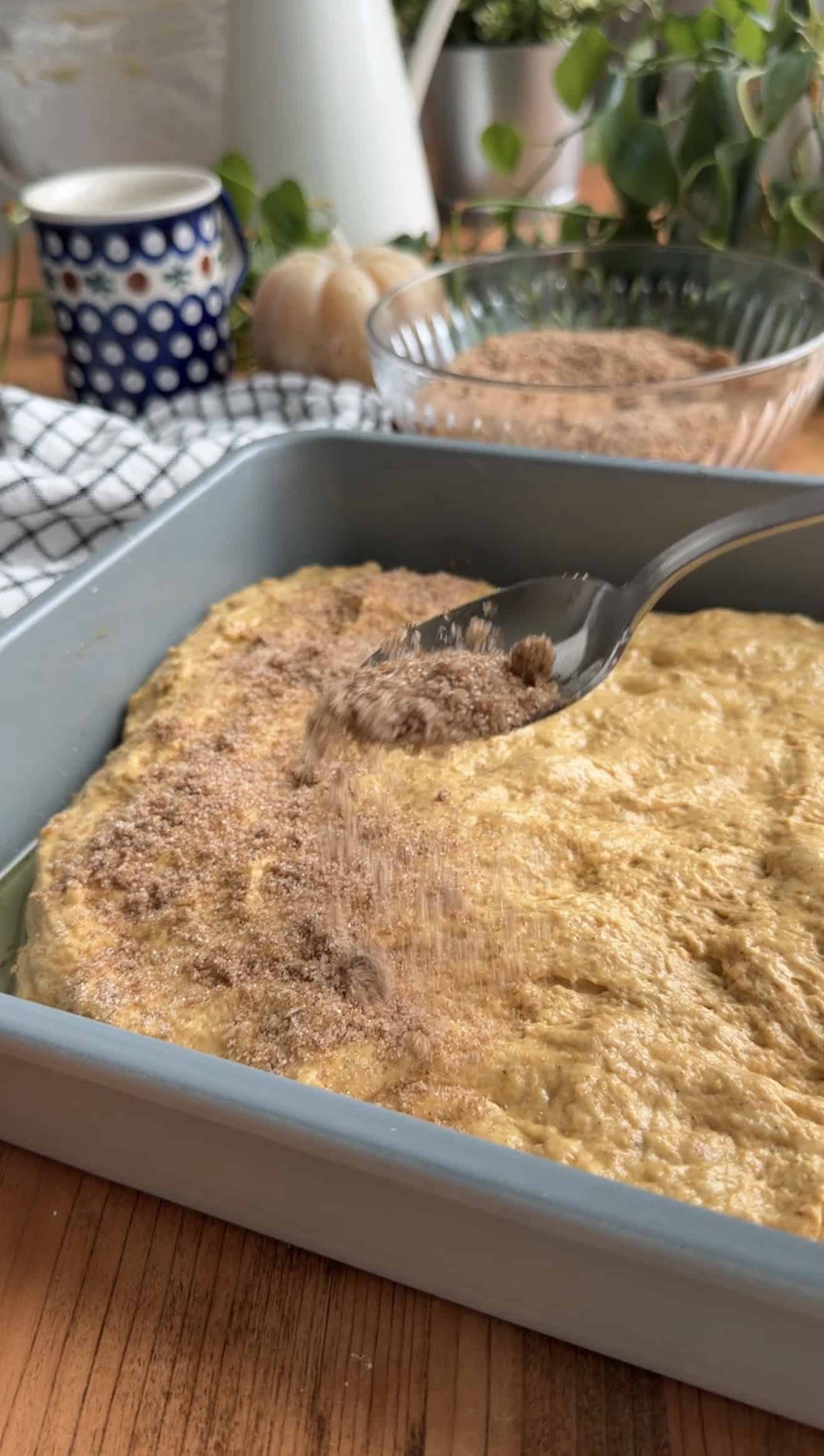 A spoon sprinkling cinnamon sugar onto orange focaccia dough in a baking dish.