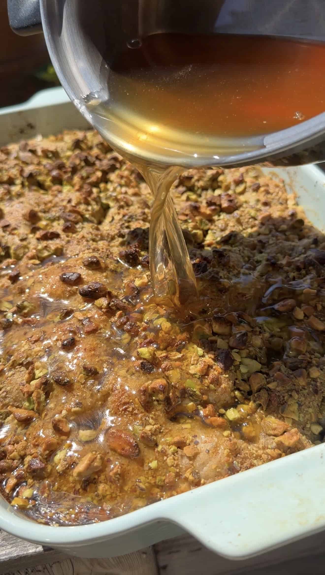 Golden syrup being poured from a metal saucepan onto baked, golden pistachio focaccia in a baking dish.