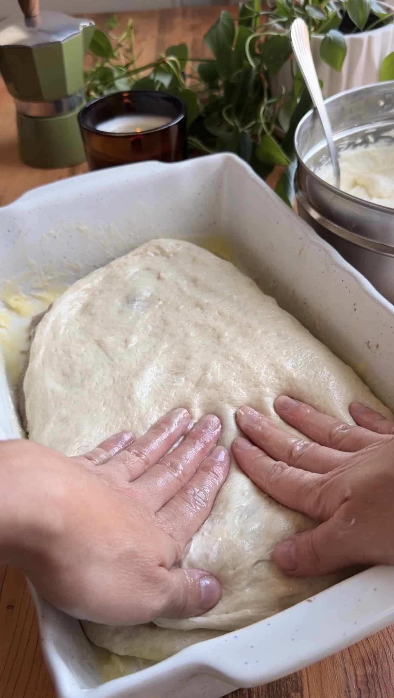 Two hands pressing focaccia dough in a white baking dish on a wooden table.