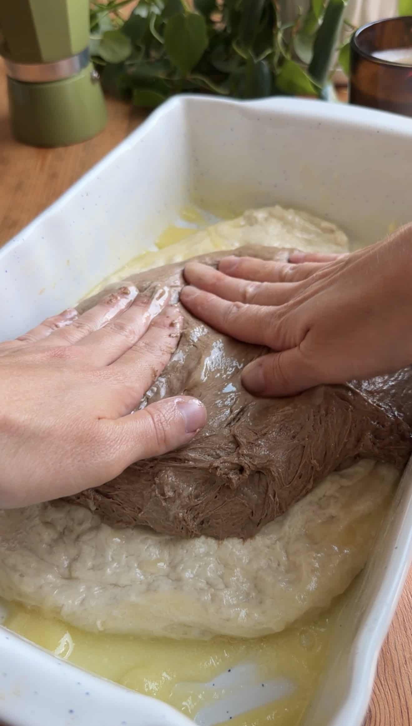 Two hands pressing out white and brown focaccia dough into a baking dish greased with melted butter.