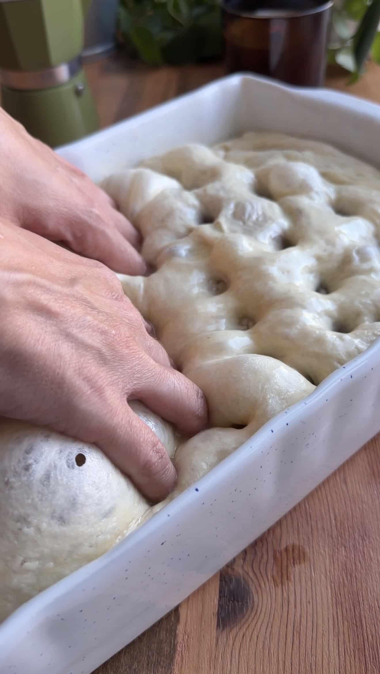 Two hands pressing indentations into puffy focaccia dough in a white baking dish.