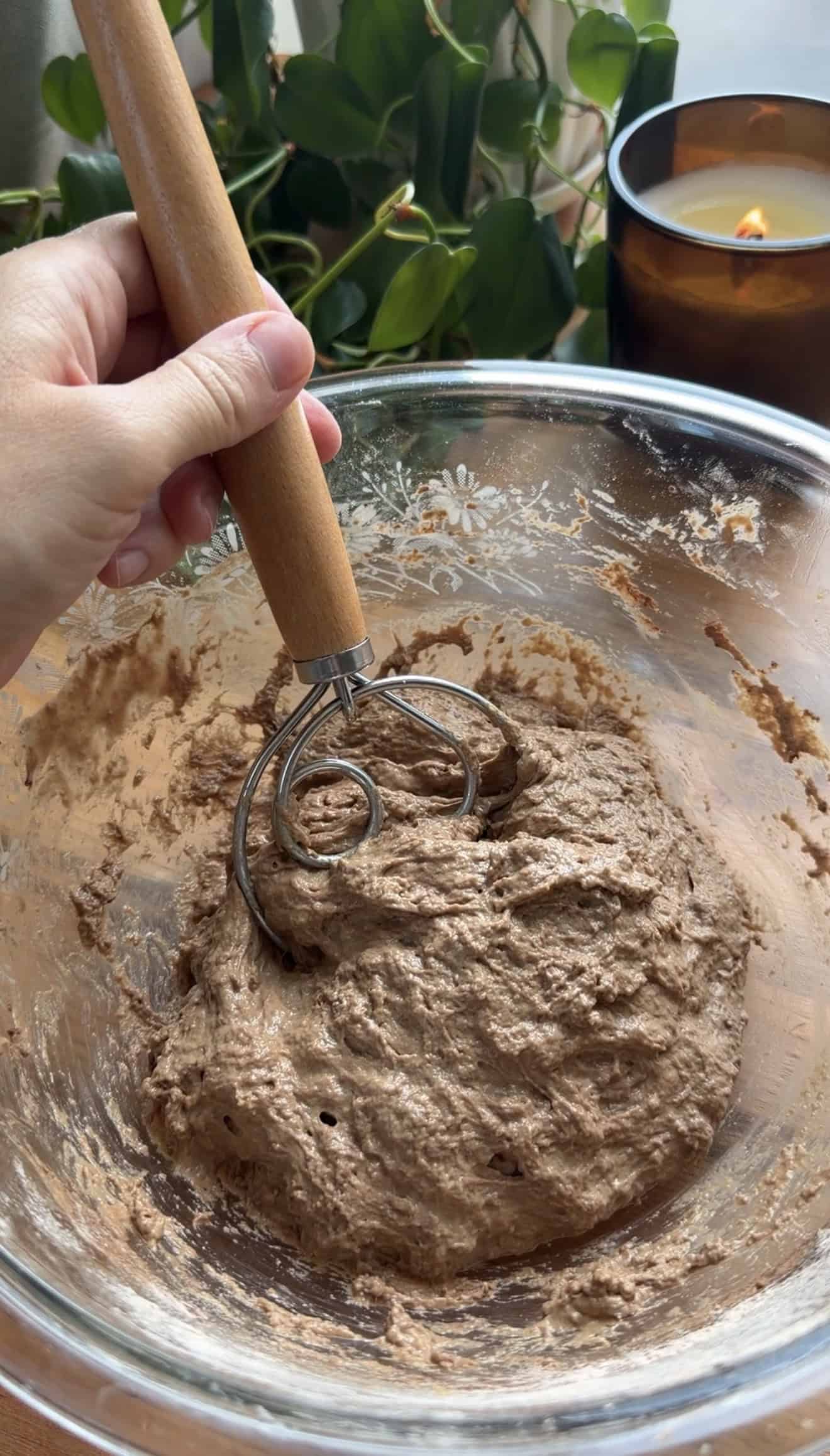 A hand mixing dark brown focaccia dough in a glass bowl with a Dutch dough whisk.
