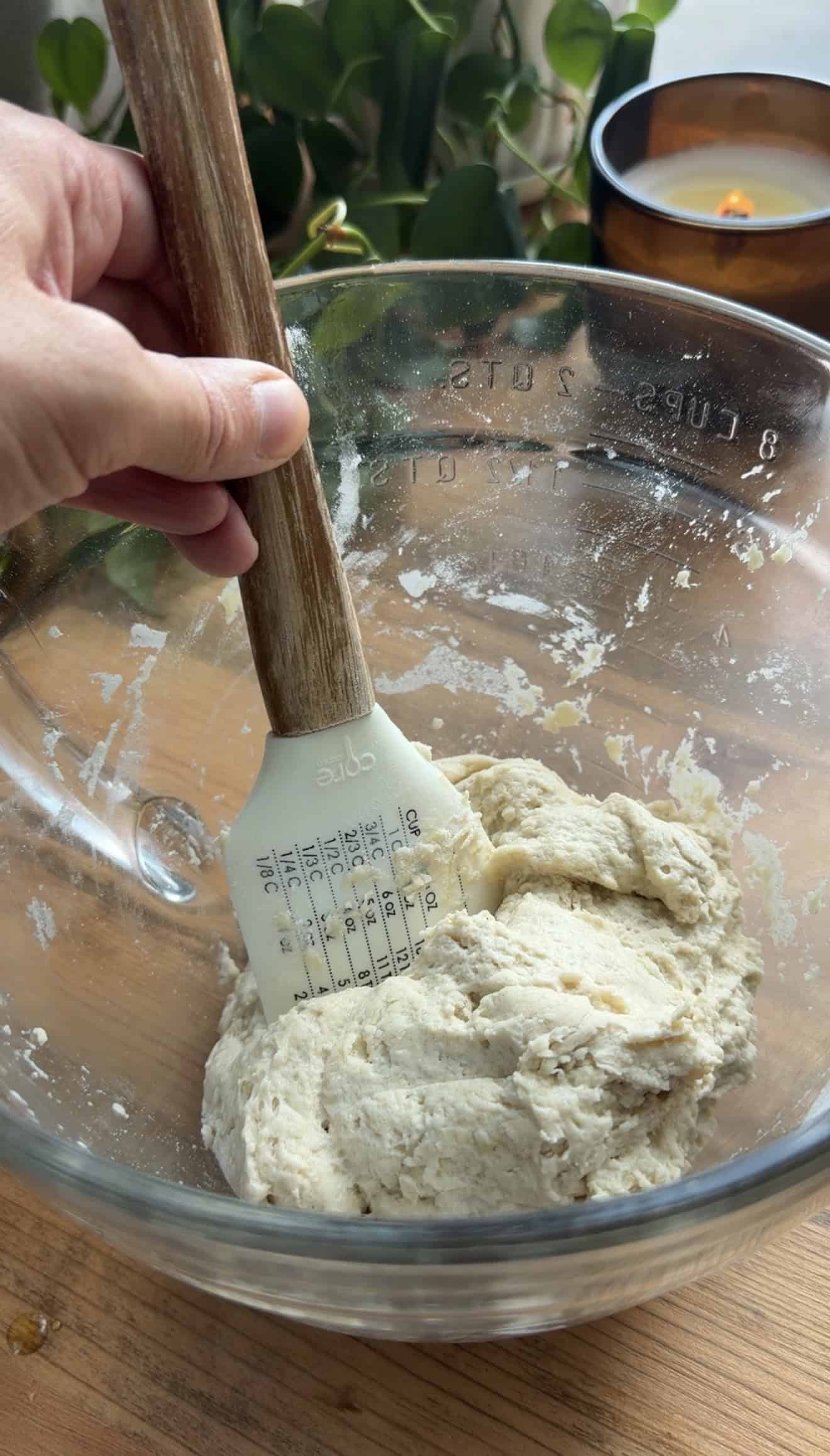 A hand mixing white bread dough in a glass mixing bowl with a silicone spatula.