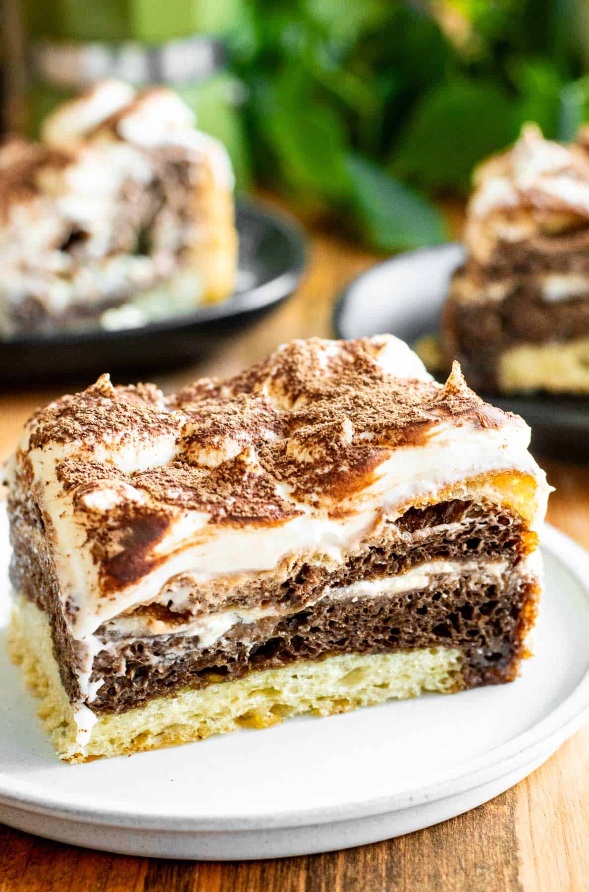 A large piece of tiramisu focaccia topped with whipped mascarpone and sifted cocoa powder next to more slices on separate plates.