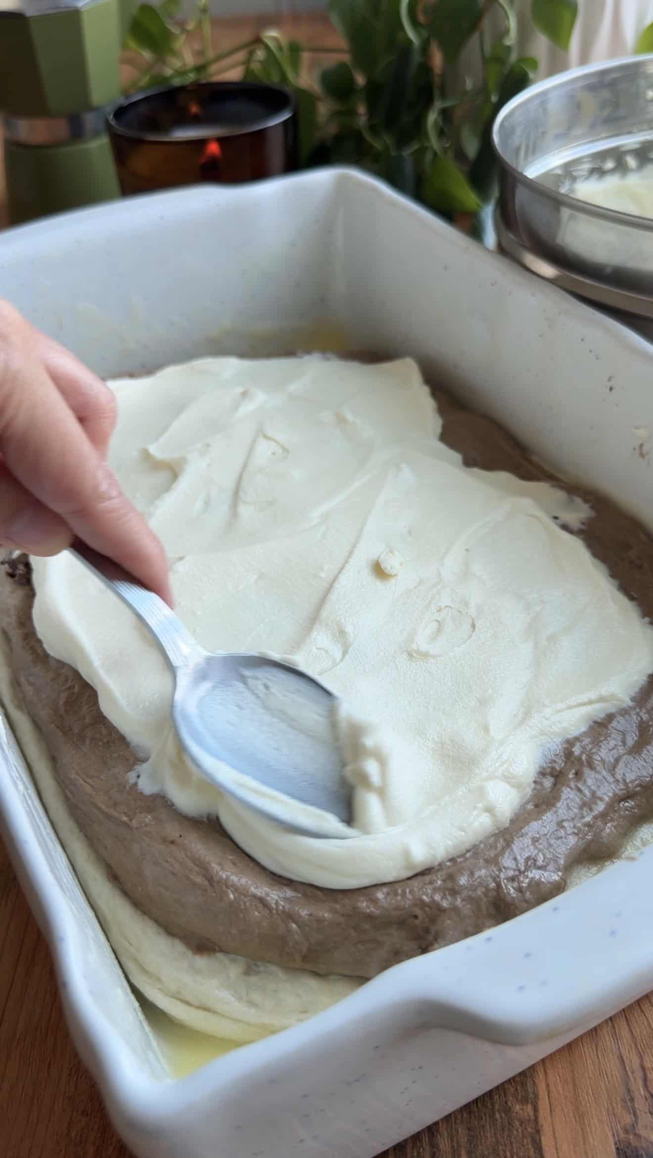 A hand spreading whipped mascarpone and cream cheese onto focaccia dough in a baking dish.