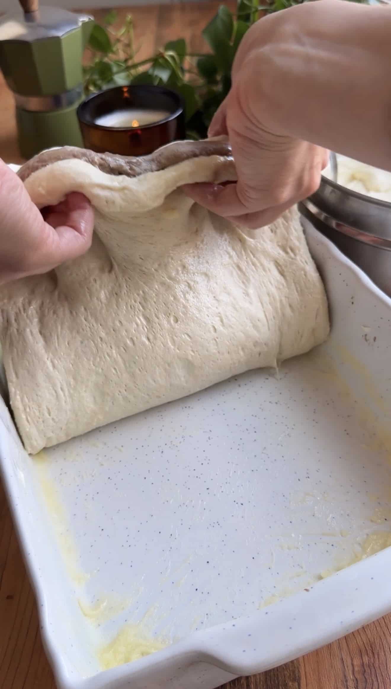 Two hands folding focaccia dough in half in a greased white baking dish.