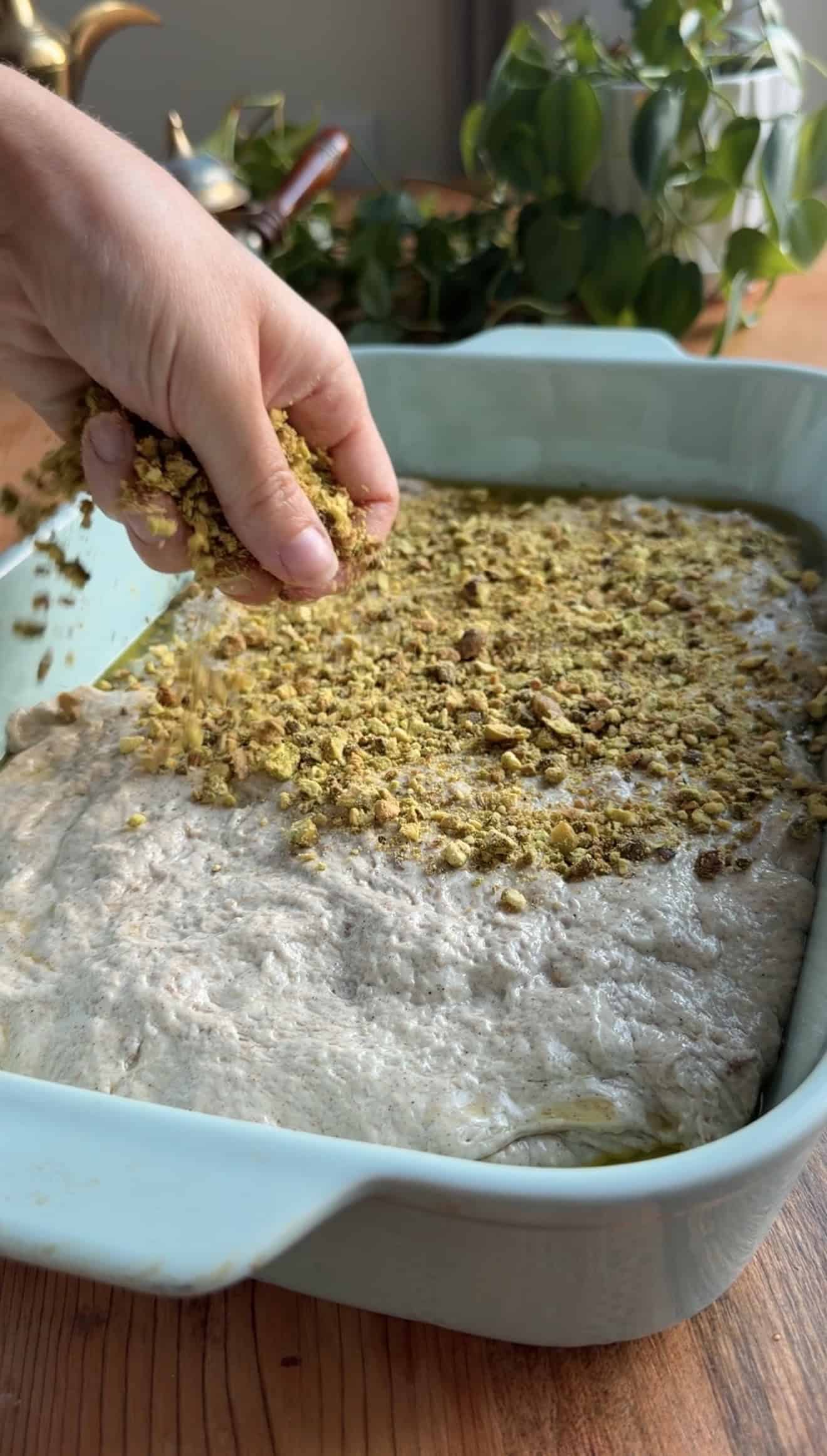 A hand sprinkling chopped pistachios on bread dough in a blue baking dish.