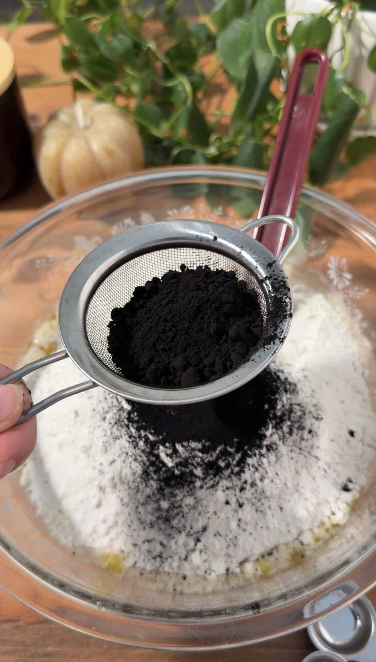 A hand sifting black cocoa powder into a glass bowl of flour.