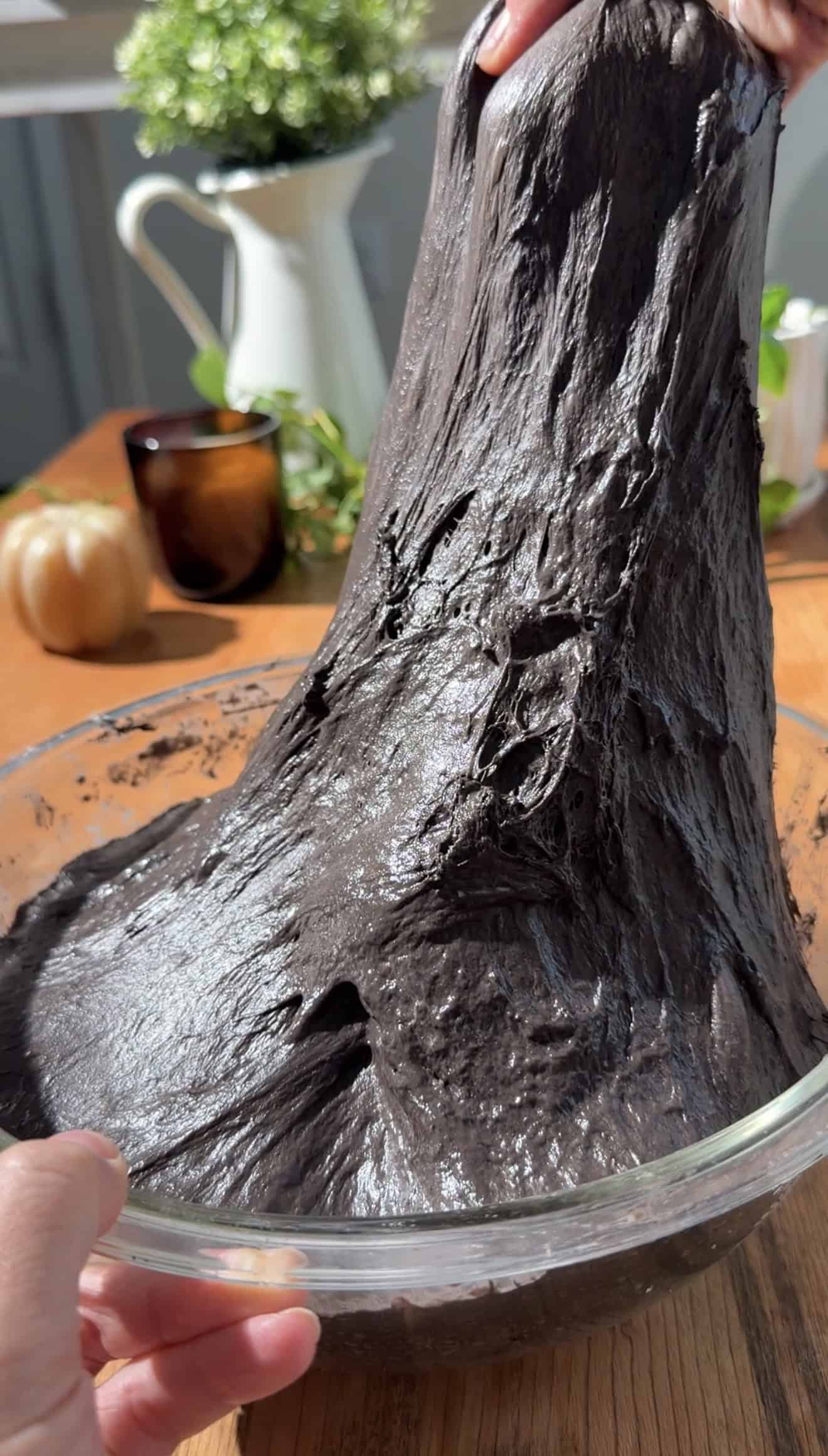 A hand stretching sticky black focaccia dough out of a glass mixing bowl.
