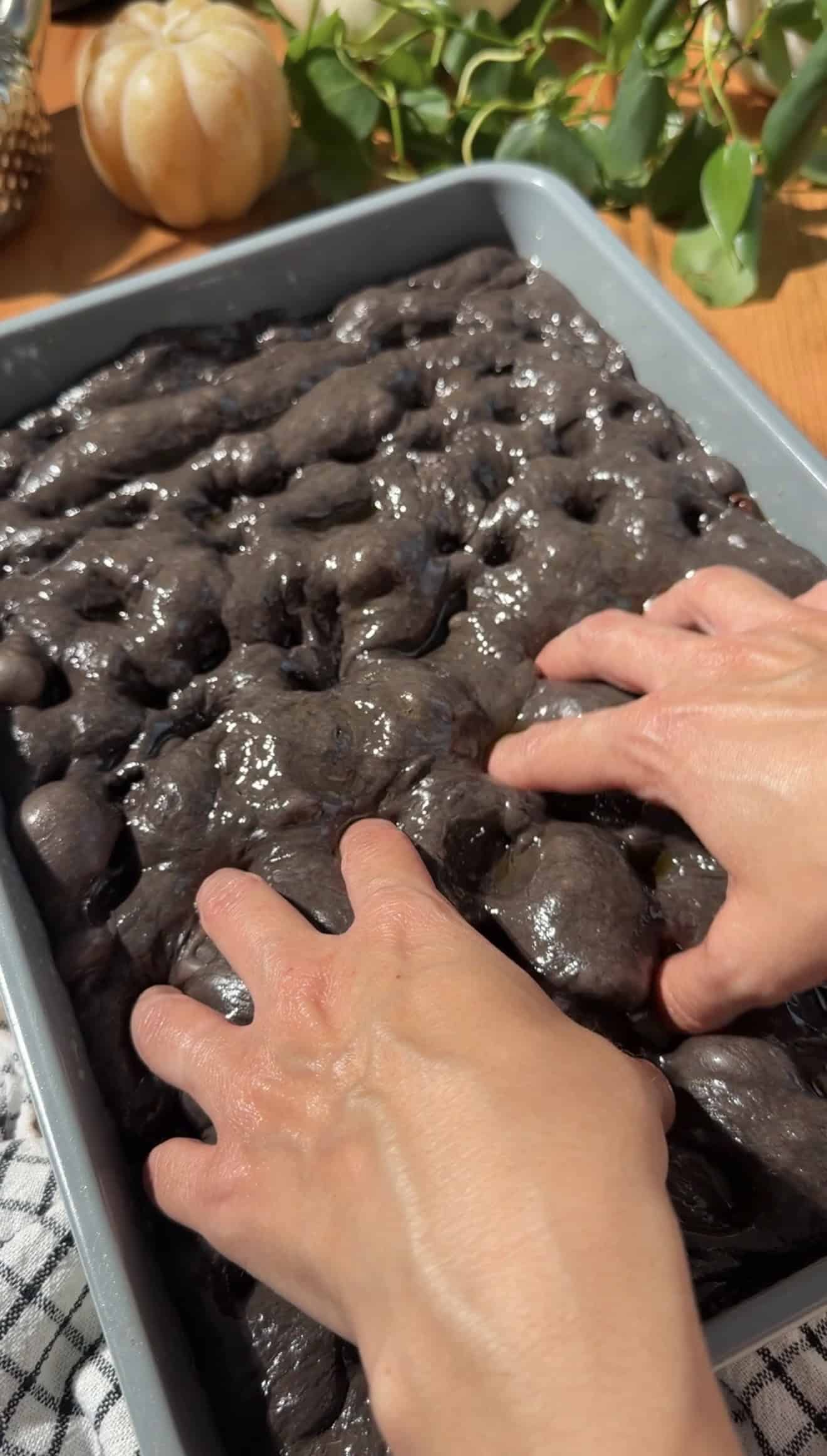 Two hands pressing dimples into black focaccia dough in a baking dish.