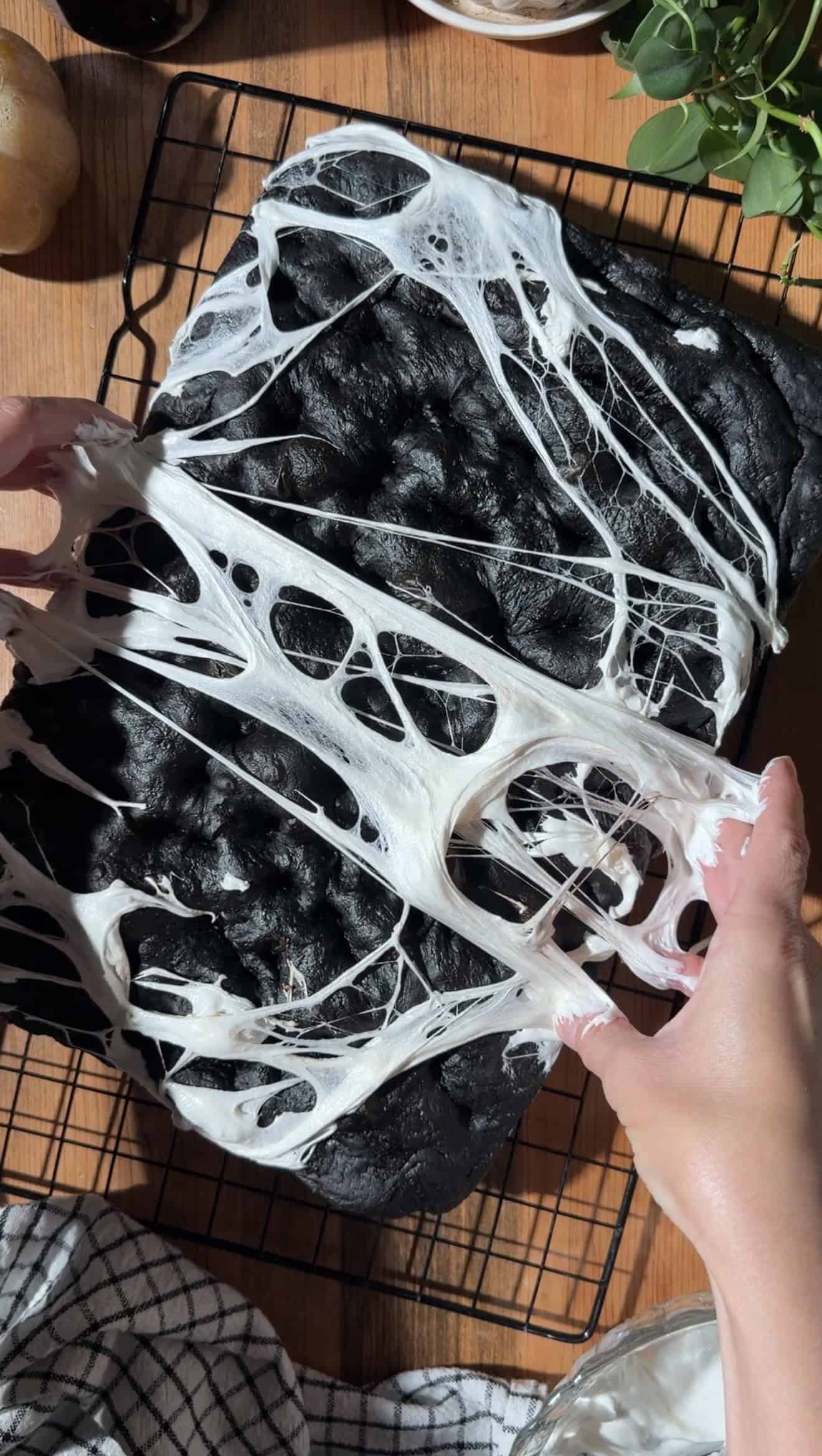 Two hands stretching melted marshmallows over black focaccia on a cooling rack.