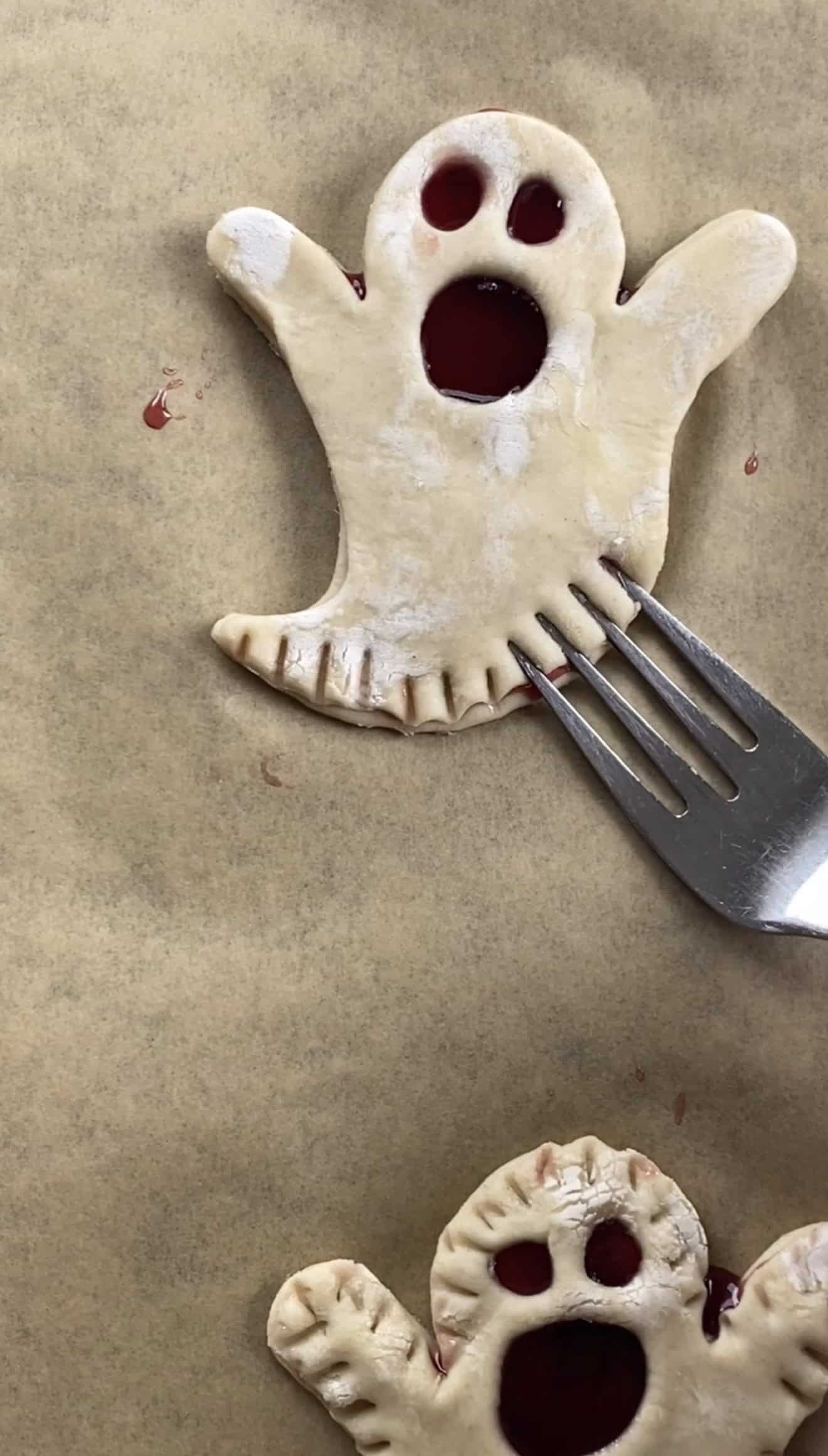 A metal fork indenting the edges of a ghost-shaped puff pastry pie that's unbaked on a baking sheet.