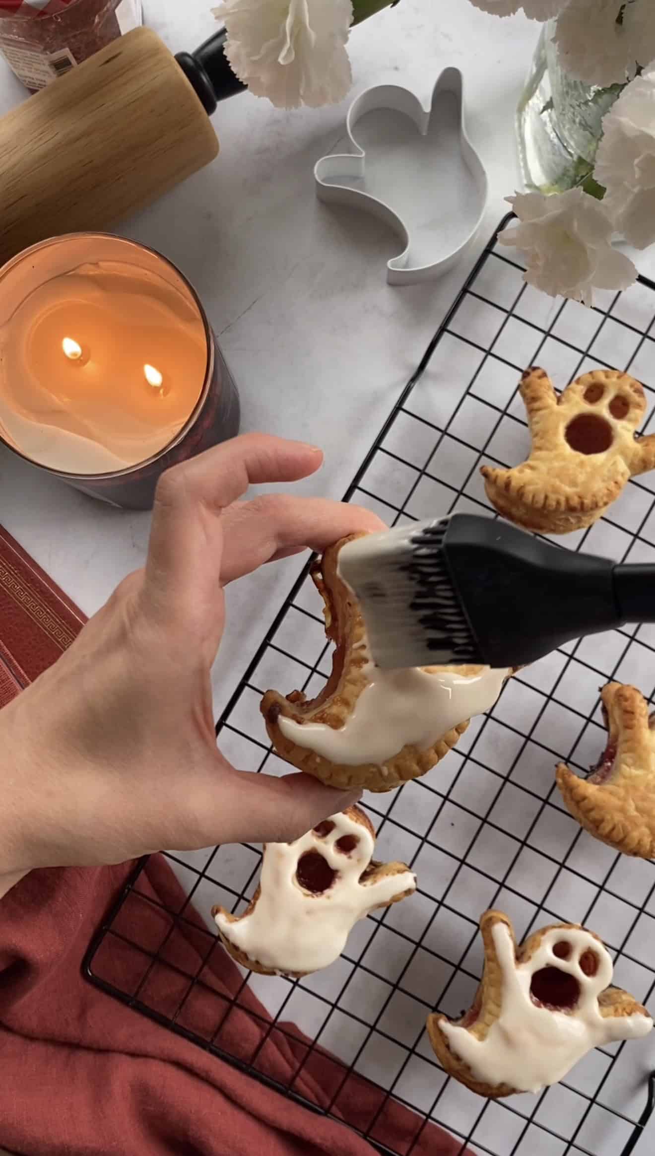 A hand brushing white glaze onto baked puff pastry pies in the shape of ghosts.