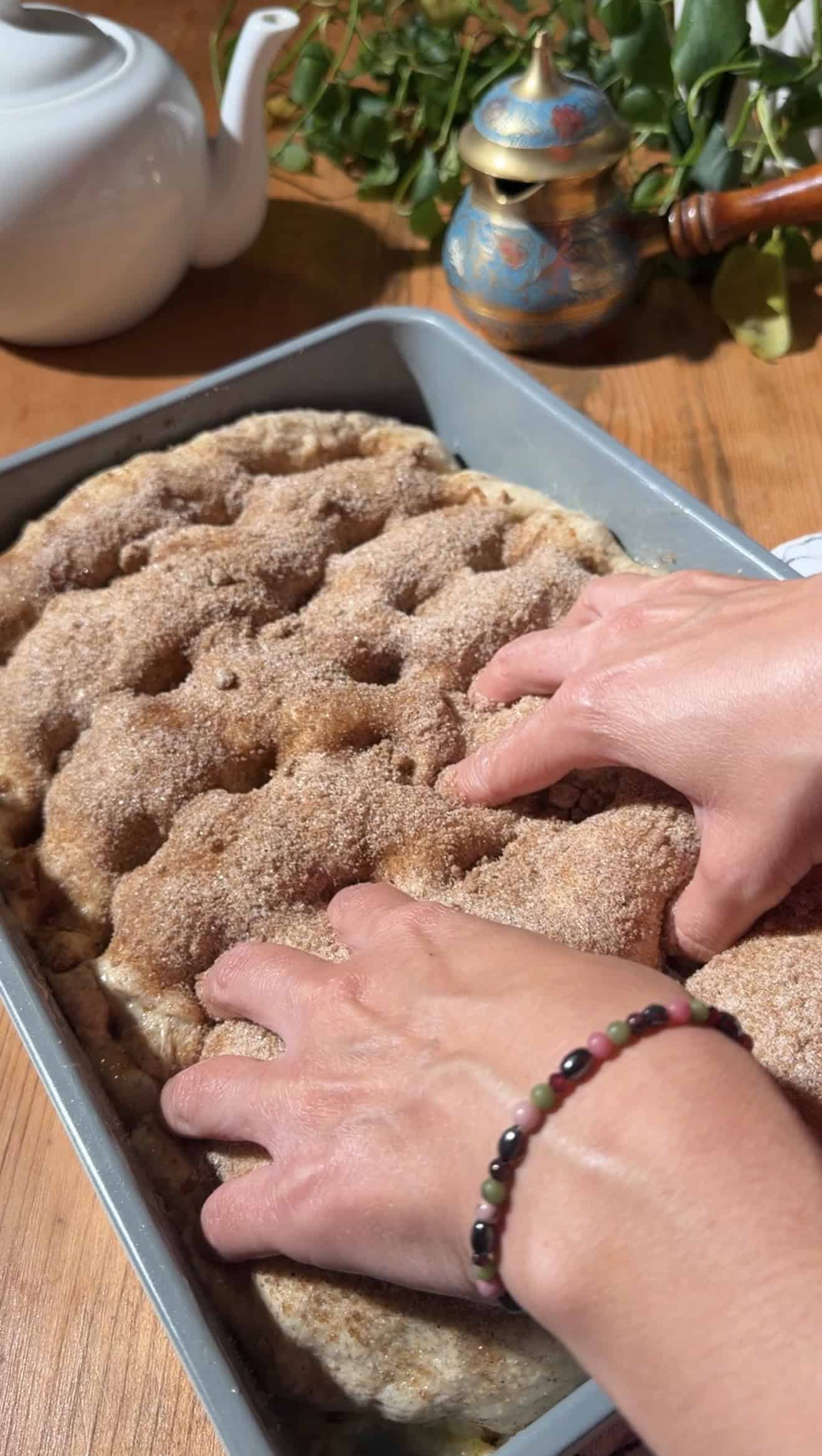 Two hands pressing dimples into the top of puffy cinnamon-sugar-coated focaccia dough in a baking dish.