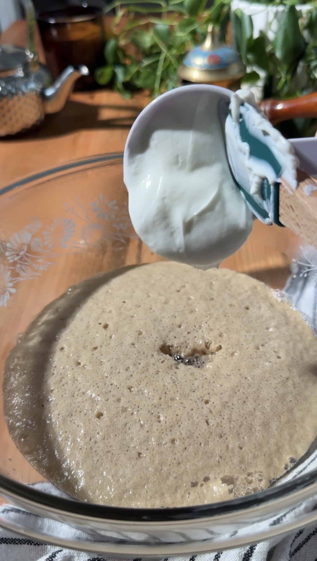 Greek yogurt being scooped out from a measuring cup into a mixing bowl of frothy yeast.