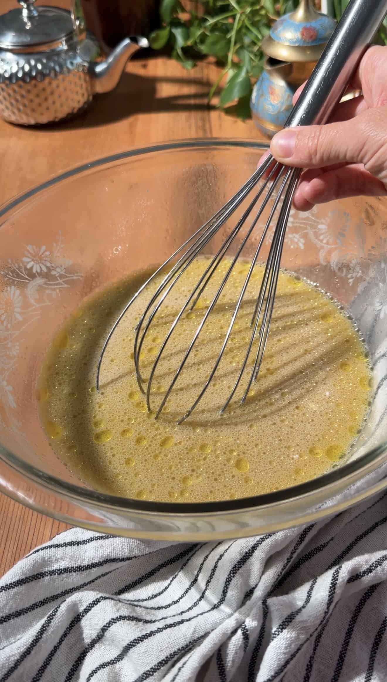 A hand whisking a yellow liquid in a glass mixing bowl on a table.