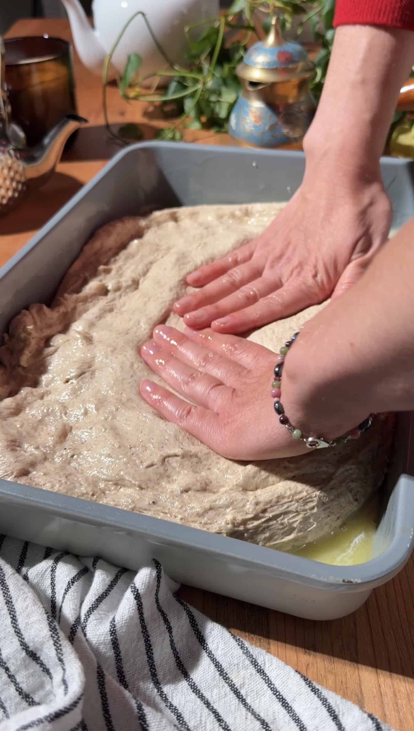 Two hands pressing focaccia dough into a metal baking dish.