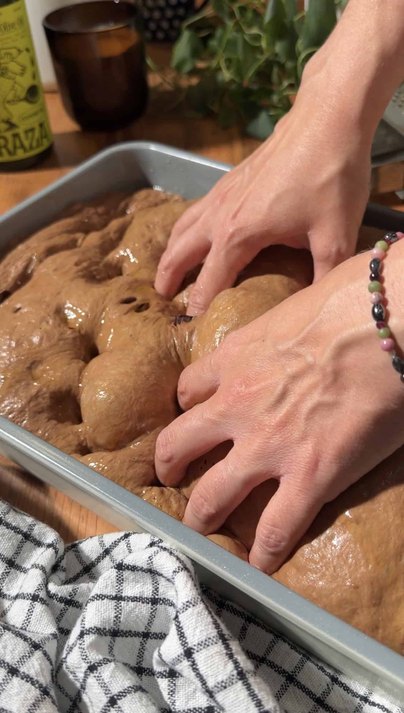 Two hands pressing dimples into puffy chocolate focaccia dough in a metal baking dish.