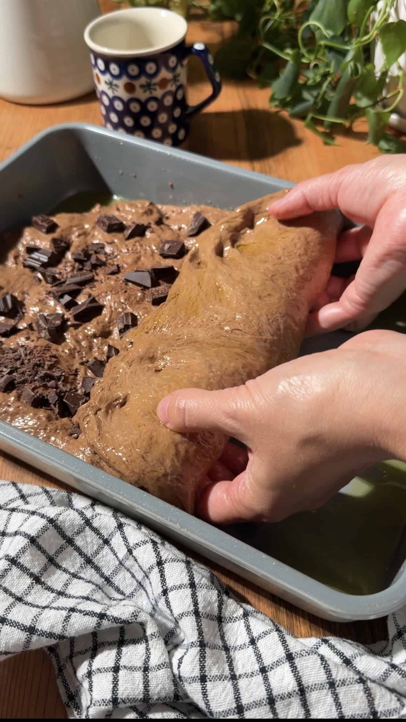 Two hands folding chopped chocolate into chocolate bread dough in a baking dish.