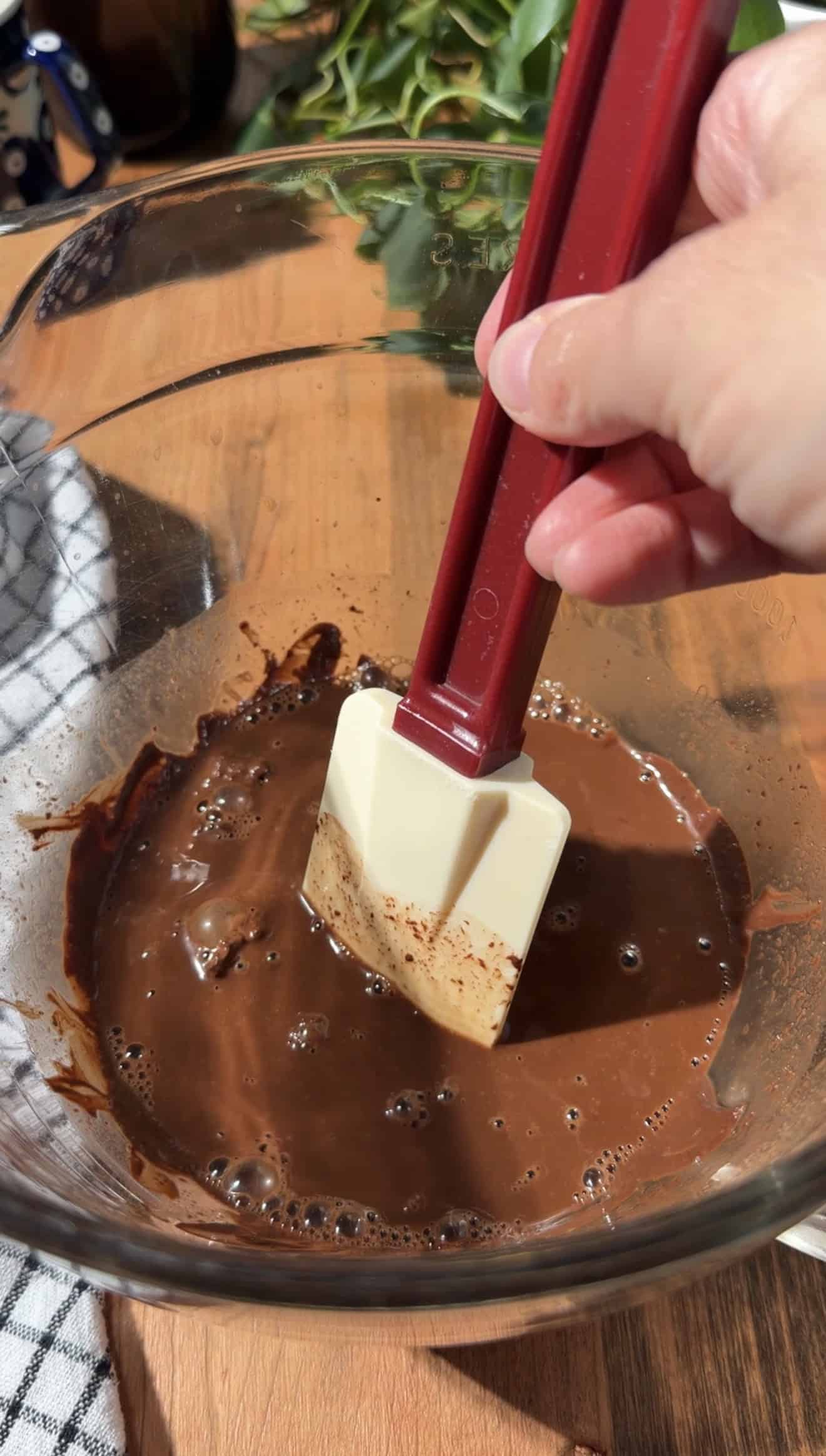 A hand stirring melted chocolate in a glass bowl with a silicone spatula.