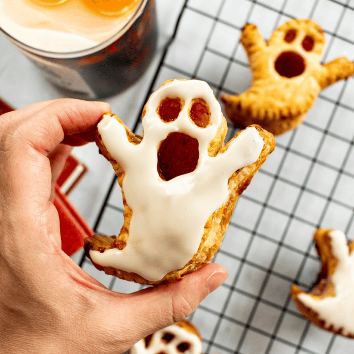 A hand holding a baked puff pastry in the shape of a ghost covered in white glaze.