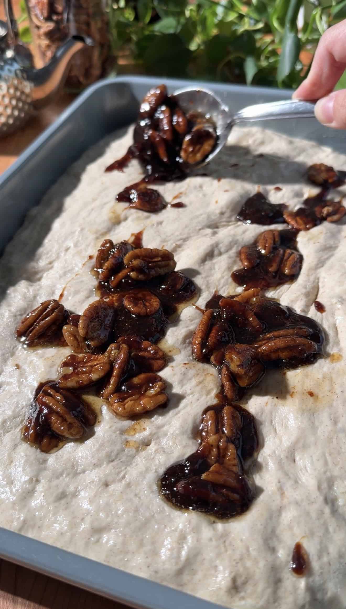 A hand spooning candied pecans and brown sugar sauce onto focaccia dough in a metal saucepan.