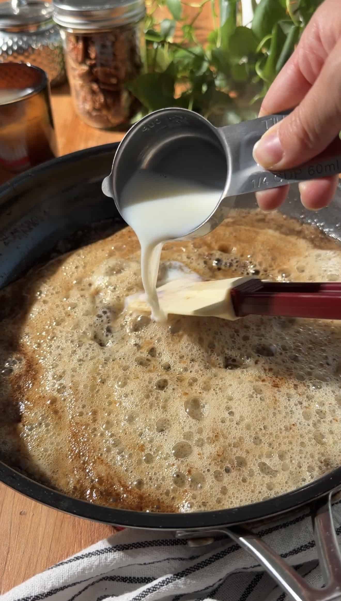A hand pouring milk from a measuring cup in a bubbling light brown liquid in a saucepan.