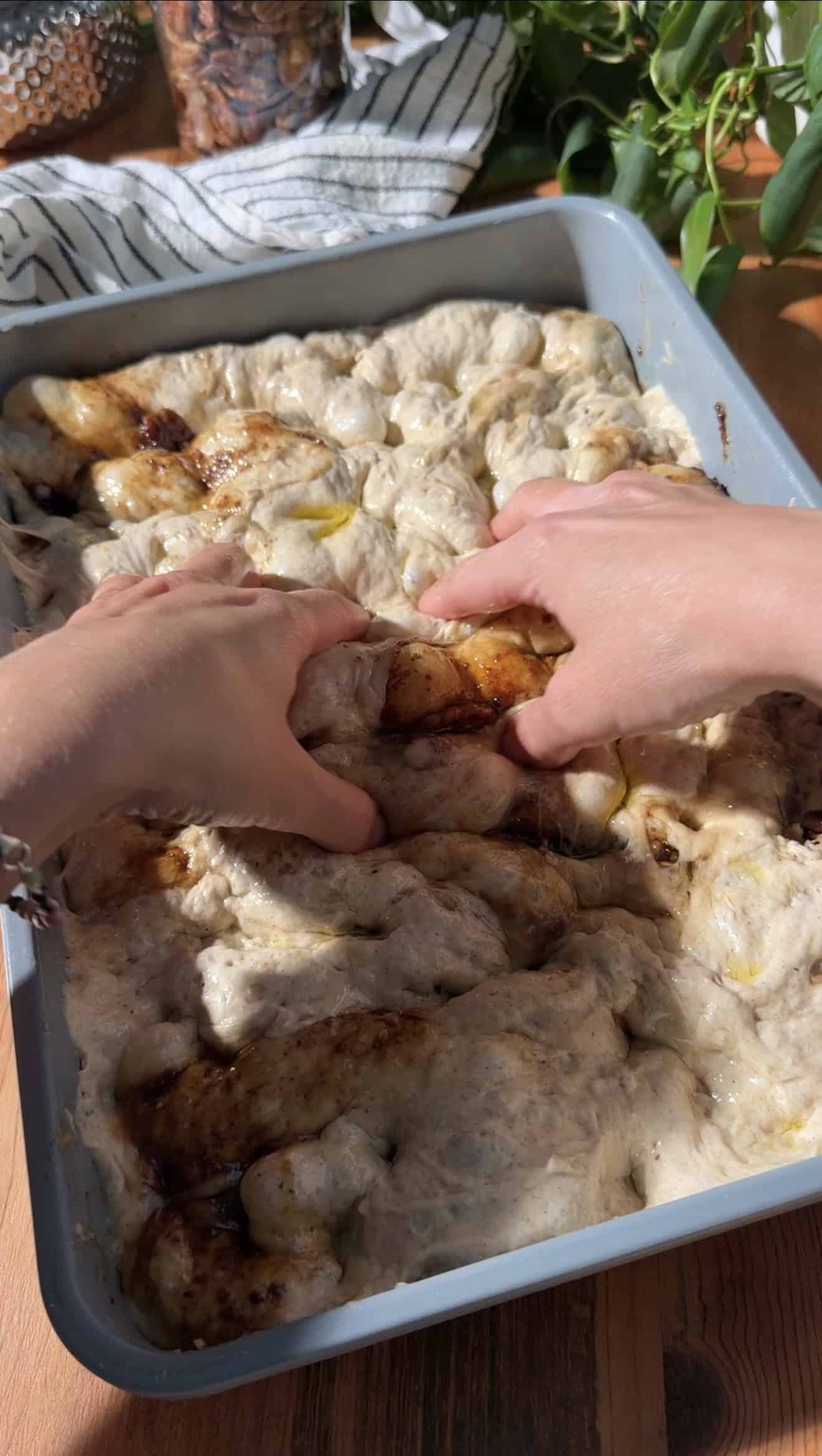 Two hands pressing dimples into focaccia dough in a metal baking dish.